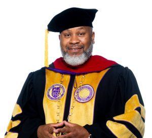 Man in academic regalia, black cap, silver beard, gold and purple medallions.
