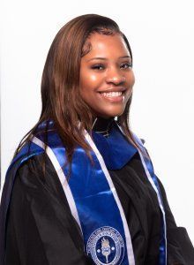 Person in graduation attire smiles, wearing a blue and white stole.