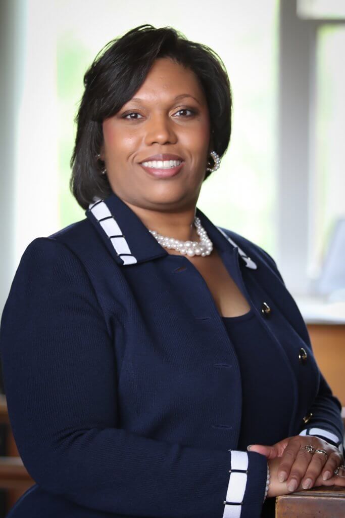 Woman in a navy blue blazer and pearl necklace smiles, posing indoors near a window and wooden furniture.
