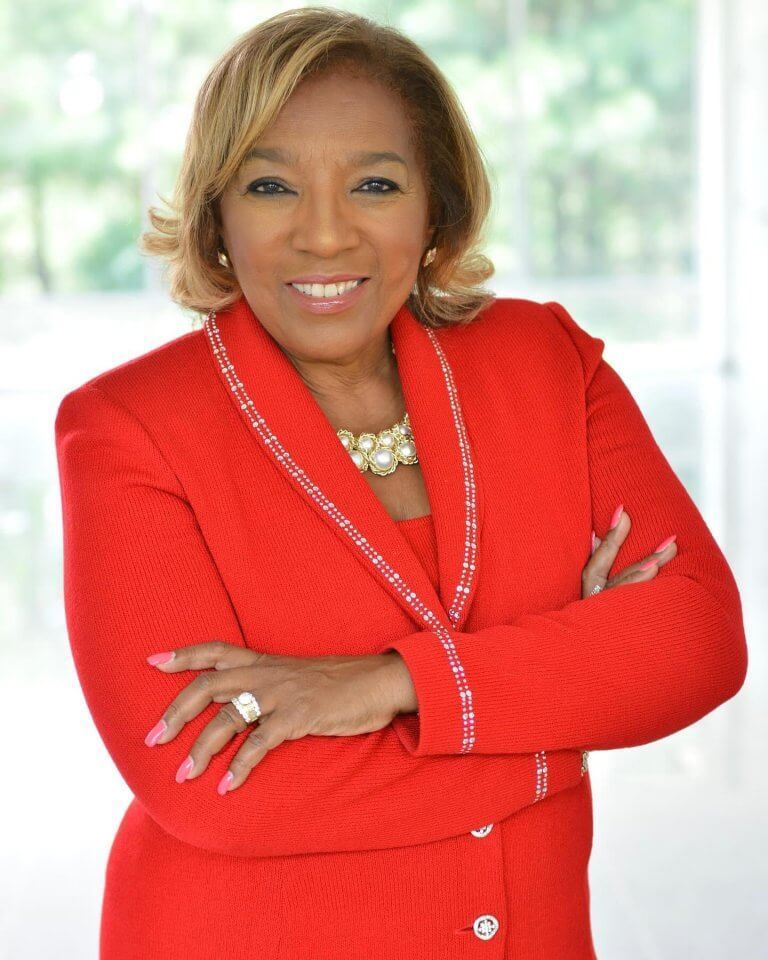 Woman in a red suit, arms crossed, smiling. Pearl necklace, ring visible. Indoor setting, bright background.