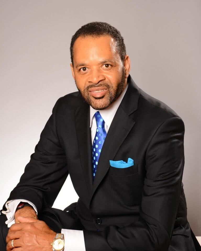 Man in black suit, blue tie, and pocket square, smiling, seated against a gray background.