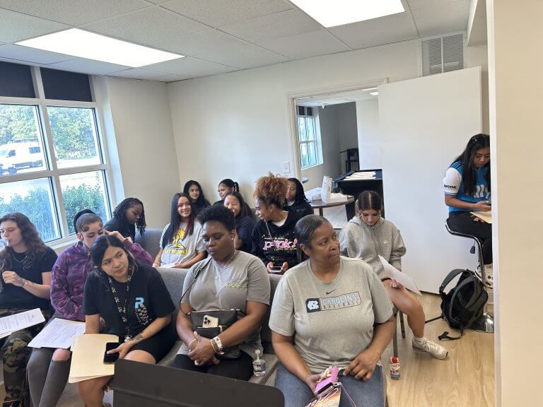 People seated in a room, some looking at notes. Daylight floods the space, and a woman writes at a table. People seated in a room, some looking at notes. Daylight floods the space, and a woman writes at a table.