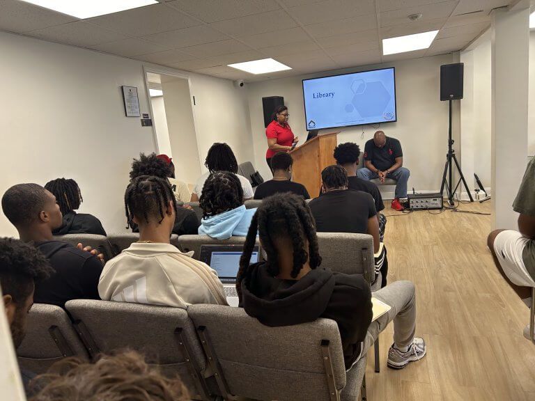 A presentation in a room. A person speaks from a podium while others listen. A screen displays text. A presentation in a room. A person speaks from a podium while others listen. A screen displays text.