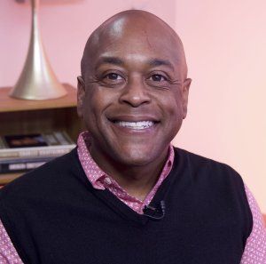 Man with a wide smile wearing a black vest over a pink collared shirt. Soft, peach-colored background.