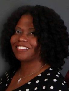 Woman with curly black hair smiles, wearing a polka dot top, and a necklace.