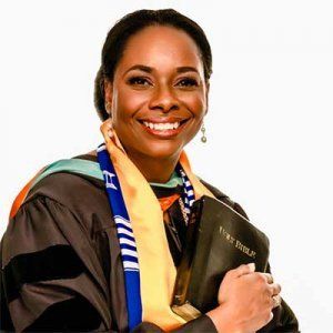 Woman in graduation robe, holding a Bible, smiling. Yellow and blue stole, against a white backdrop.