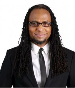 Man with long dreadlocks wearing glasses, suit, and tie, smiling.