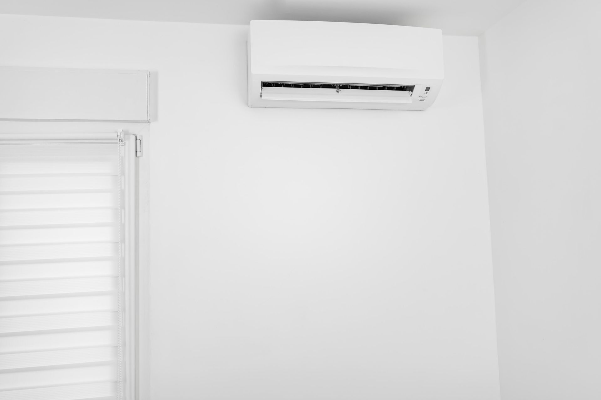White air conditioner on a white wall, above a window with white blinds.
