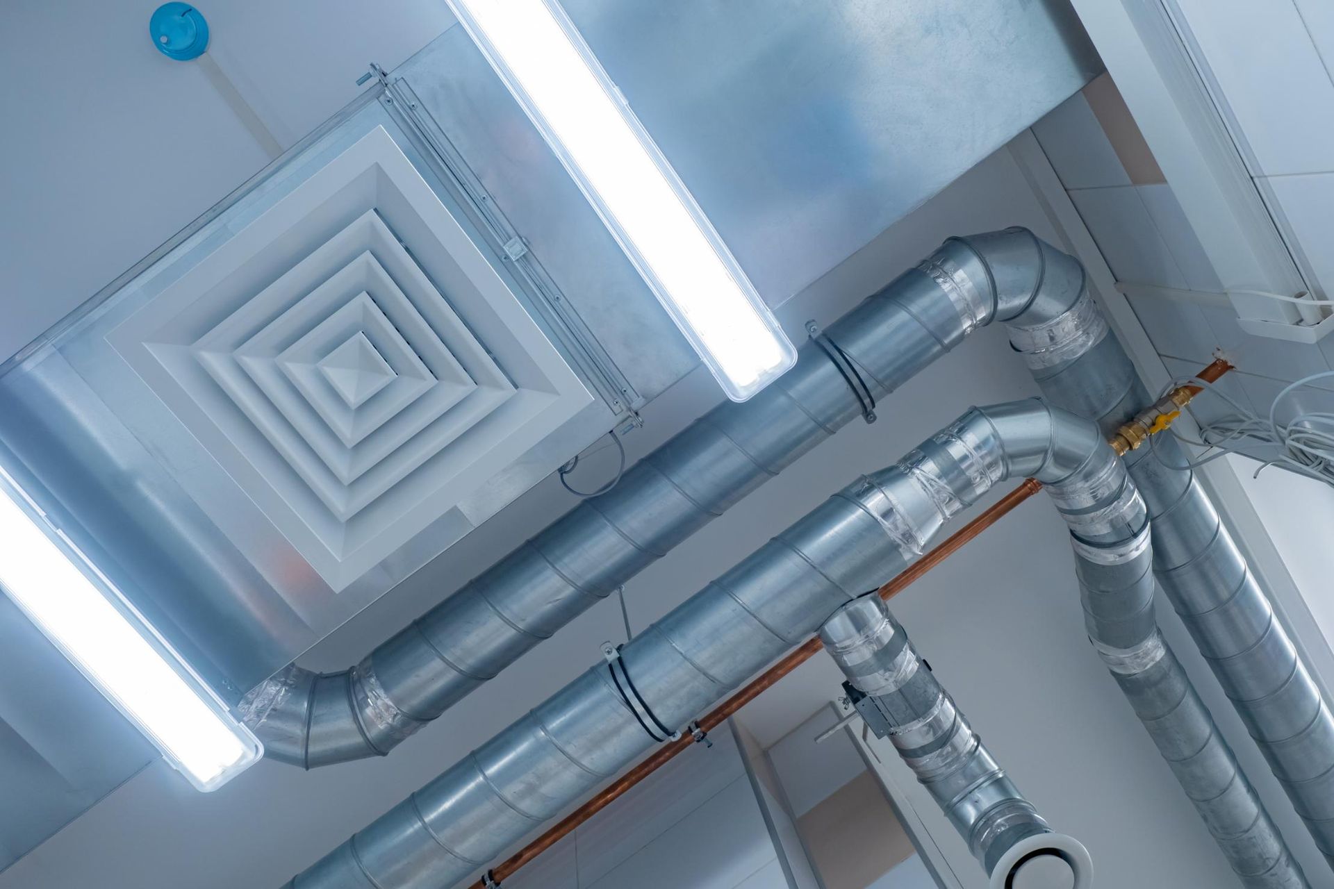 Overhead view of ductwork, vent, and fluorescent lights on a white ceiling.
