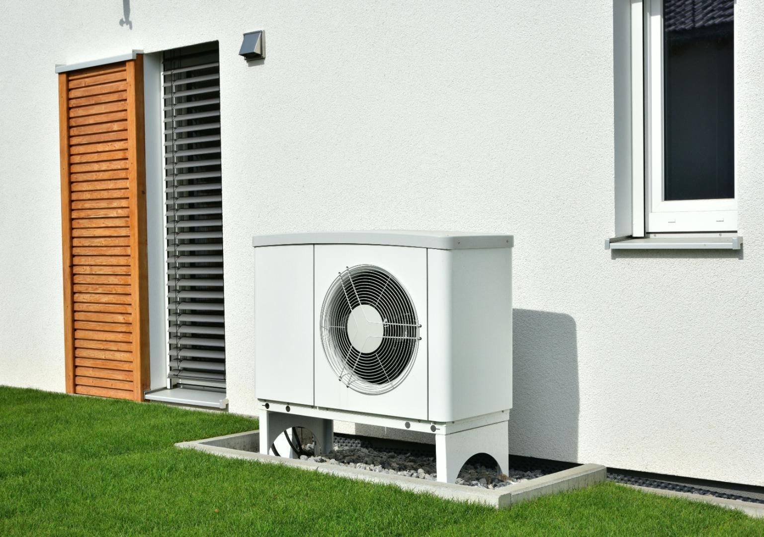 Heat pump unit on a concrete pad next to a white building with a window and wooden shutter.