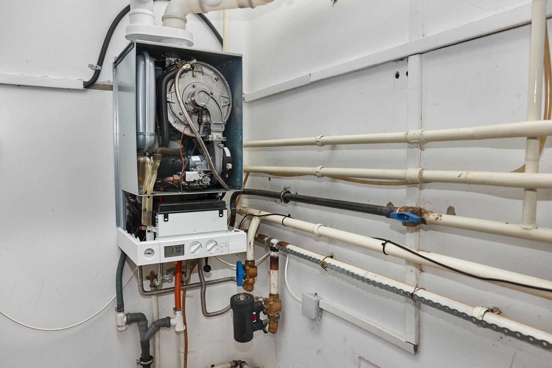 A wall-mounted gas boiler with open front, surrounded by pipes and wiring, in a white utility room.