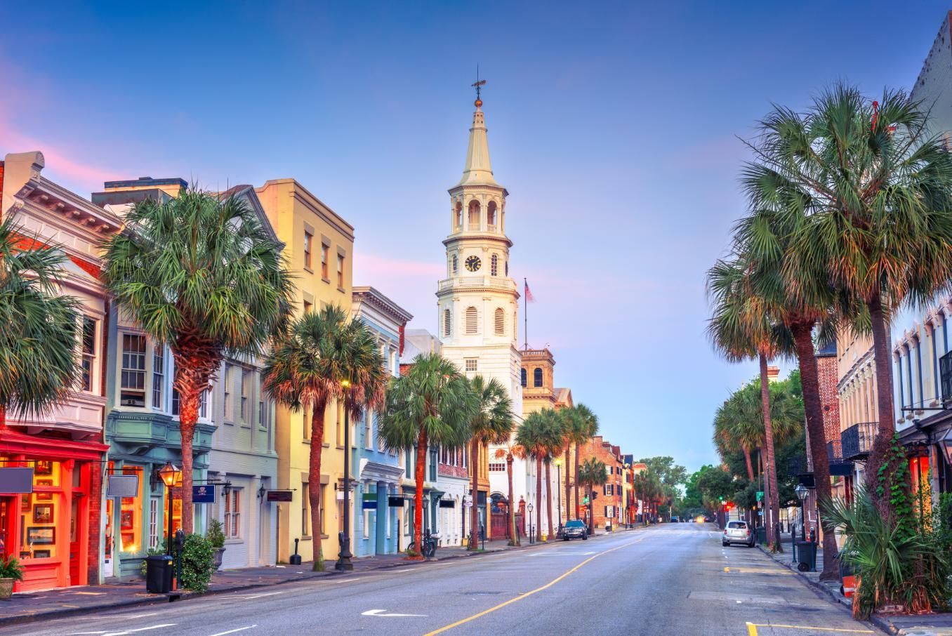 Street view of Charleston, South Carolina, with colorful buildings, palm trees, and a church steeple.