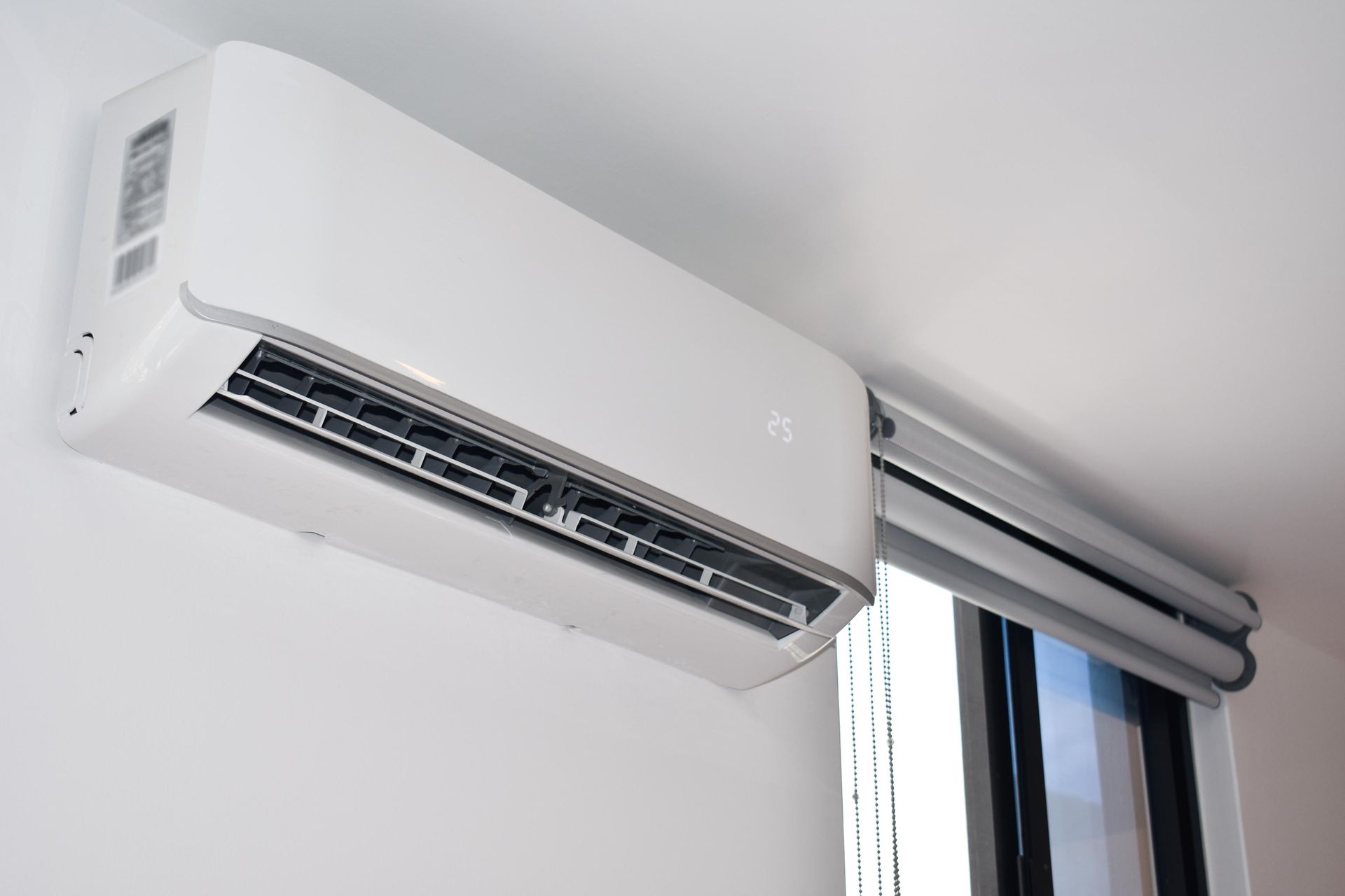 White split-type air conditioner mounted on a white wall above a window with a blind.
