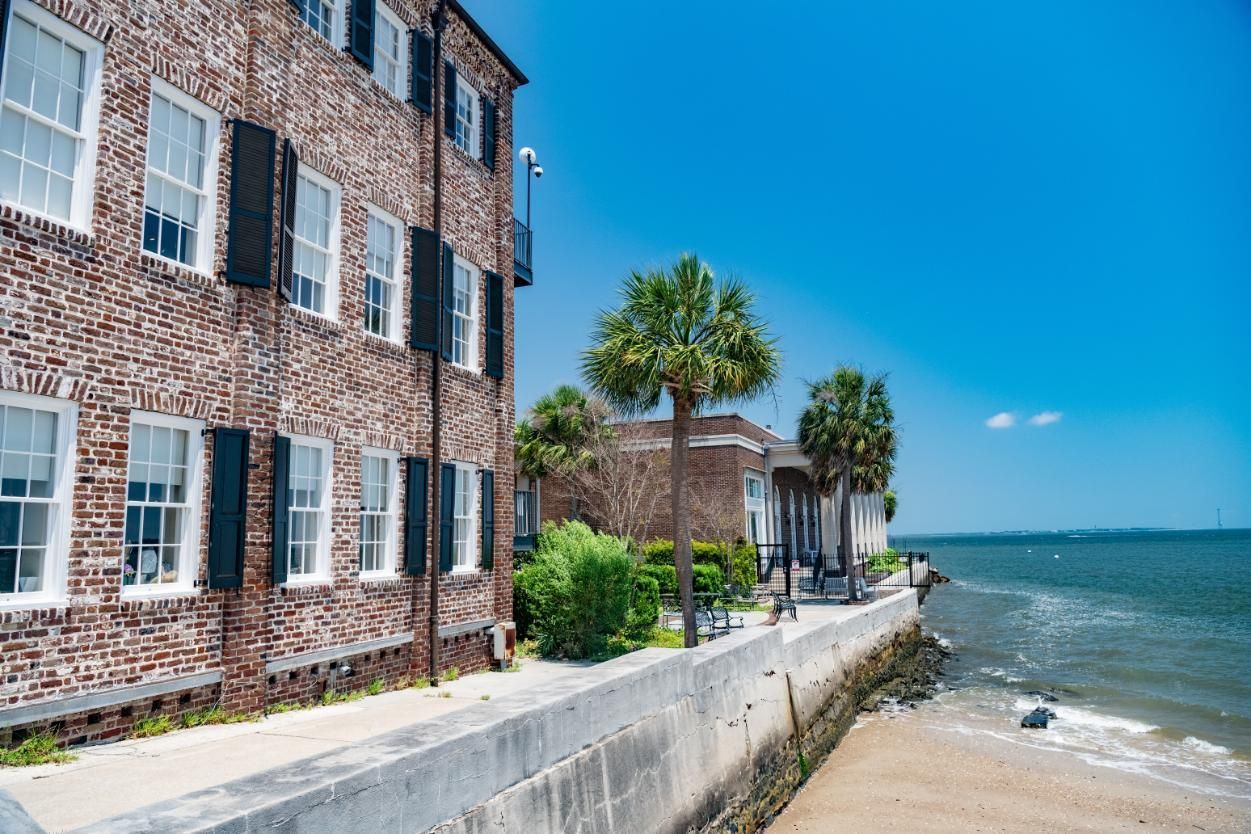 Brick building by the ocean with white windows, black shutters, and palm trees under a bright blue sky.