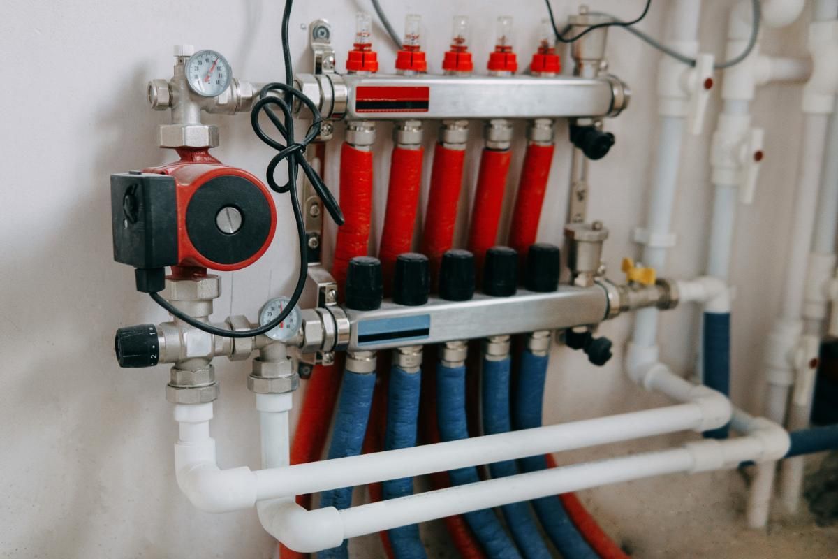 A hydronic radiant floor heating manifold with red and blue tubes, gauges, and a pump against a white wall.