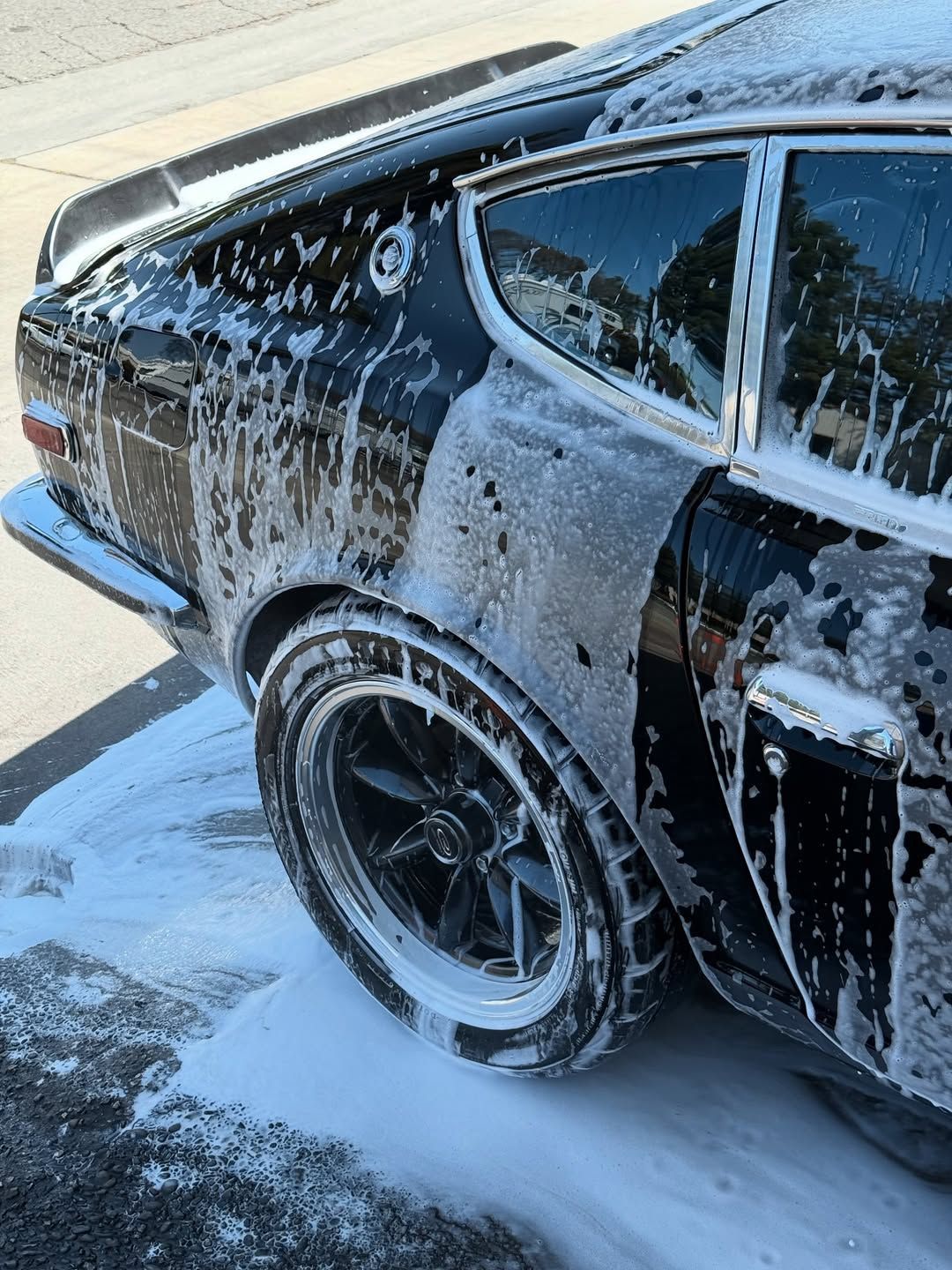 A black vintage sports car covered in thick white soap suds, parked outdoors during a car wash.