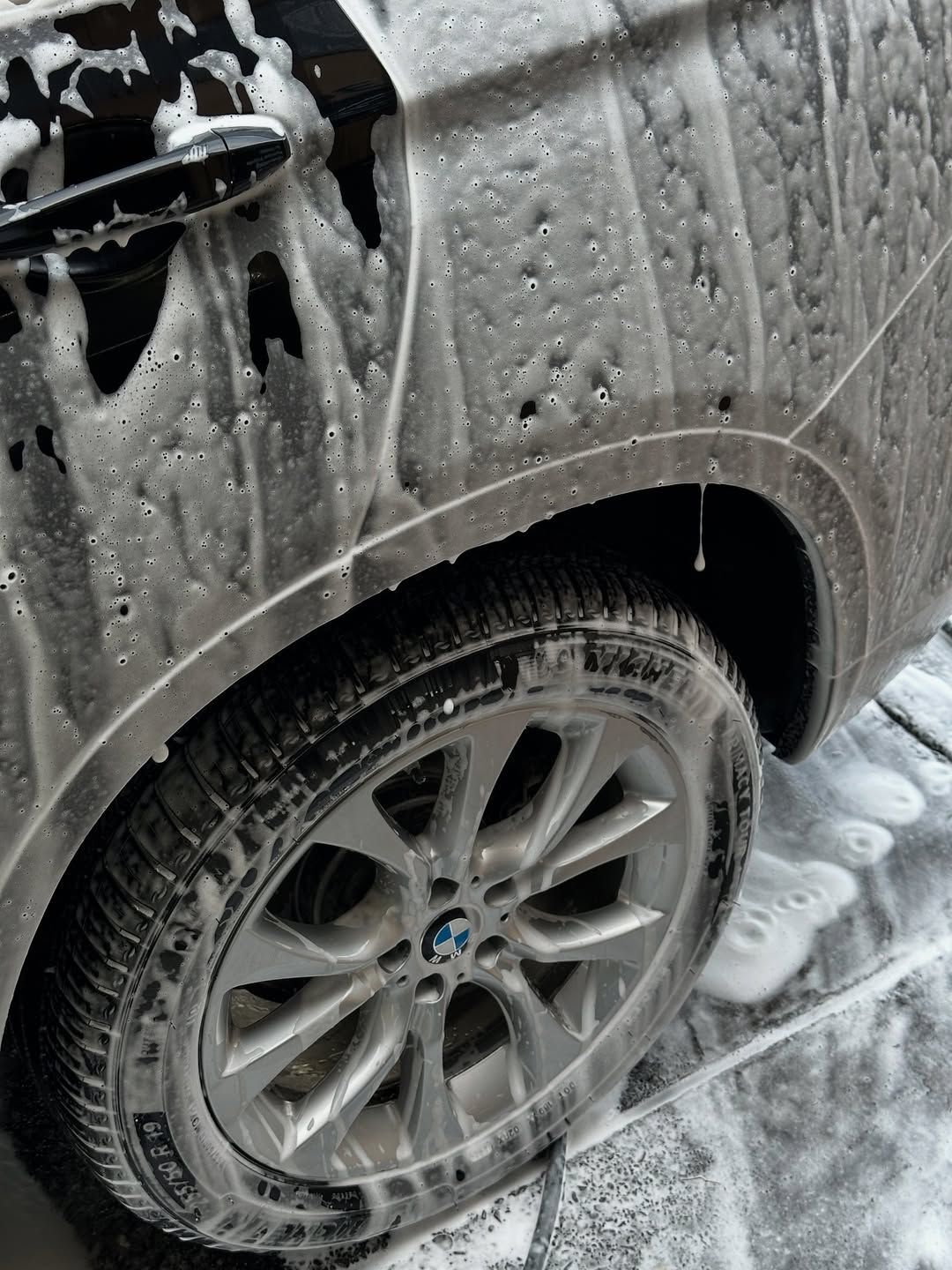 Close-up of a soapy, white-foamed BMW car wheel and side panel being washed.