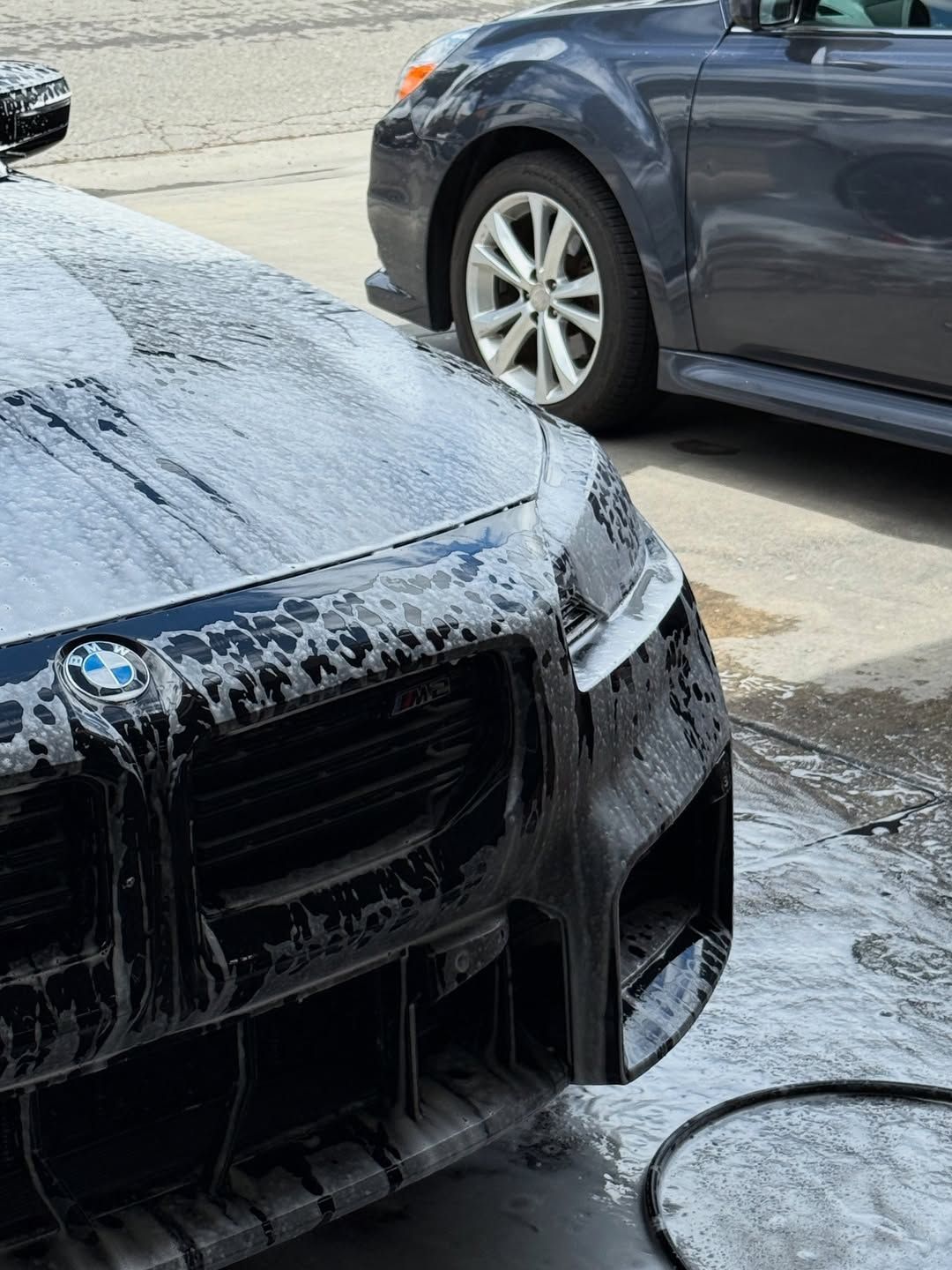 A BMW front bumper covered in soapy suds during a car wash, with a gray car parked nearby in the background.