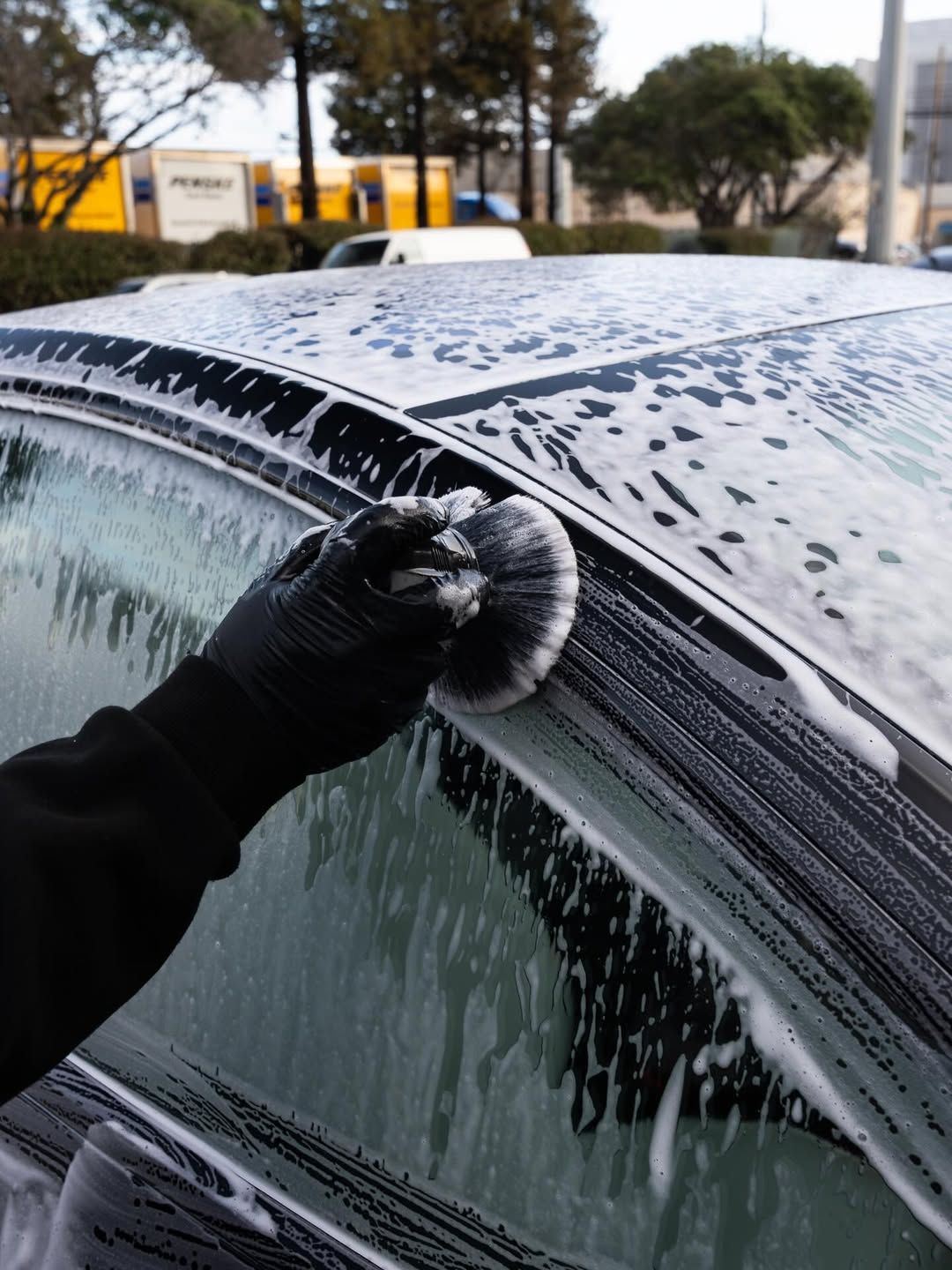 A person wearing black gloves uses a round brush to clean soapy residue from the window trim of a car.