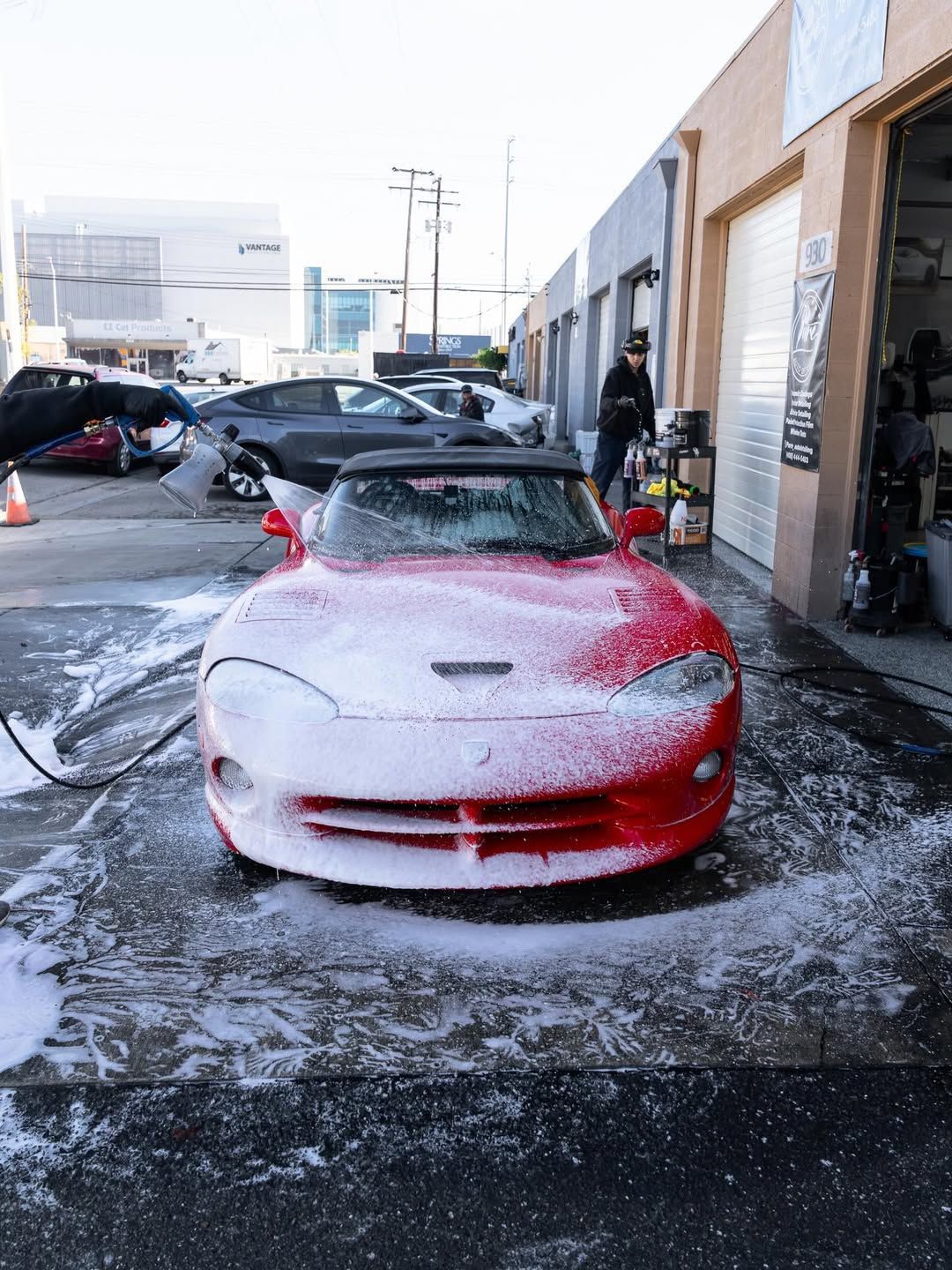 A red sports car covered in thick white soap suds parked in front of a garage, with a person working in the background.