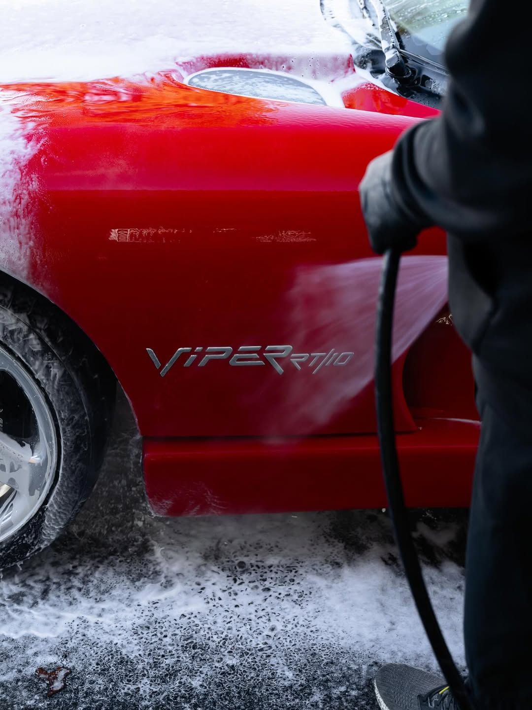 A person in black gloves power-washing soap off the side of a red Dodge Viper, clearly showing the car’s emblem.