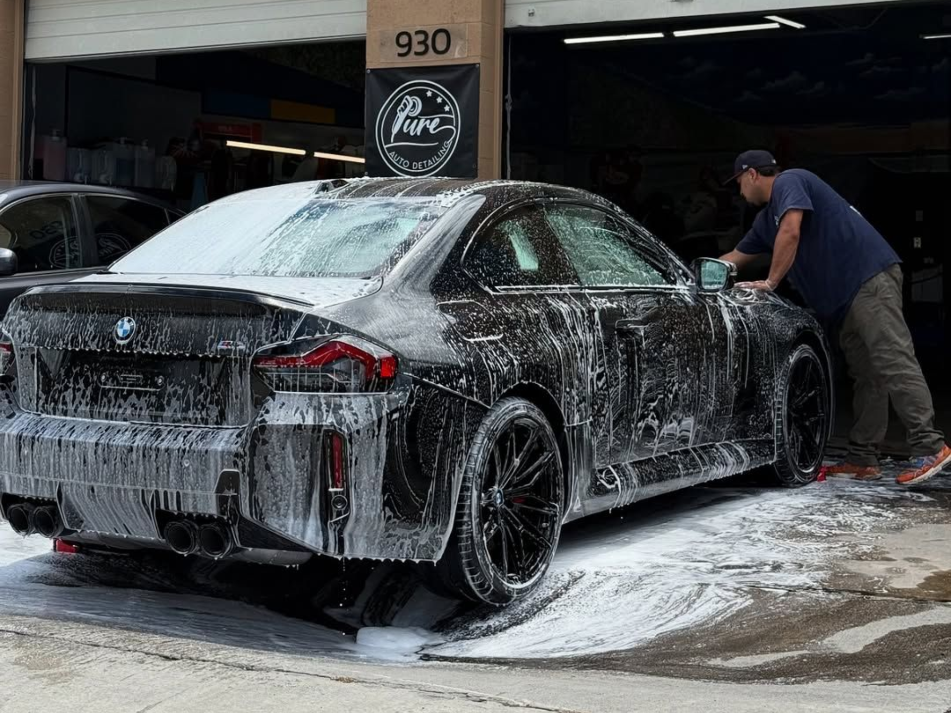 A dark-colored BMW covered in white soap suds is being washed by a person at a car detailing shop.