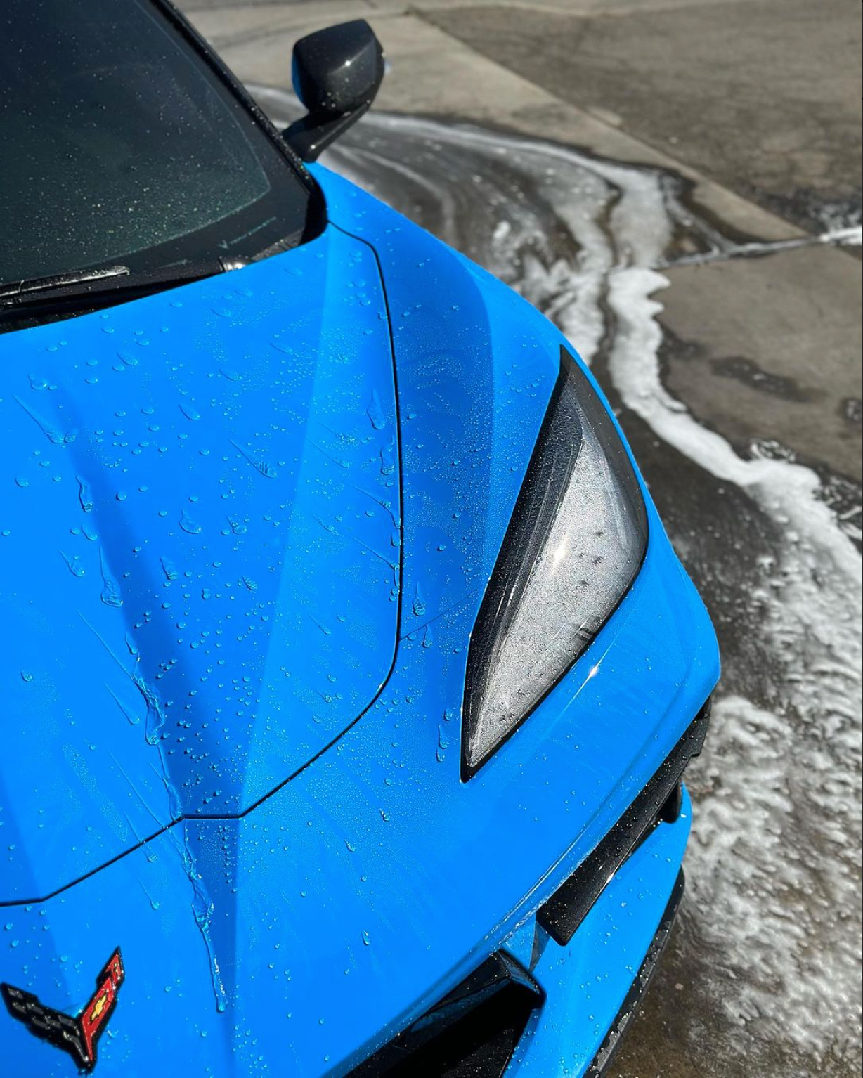 Bright blue Chevrolet Corvette C8 being washed, featuring raindrops on the hood and soapy runoff on the ground.