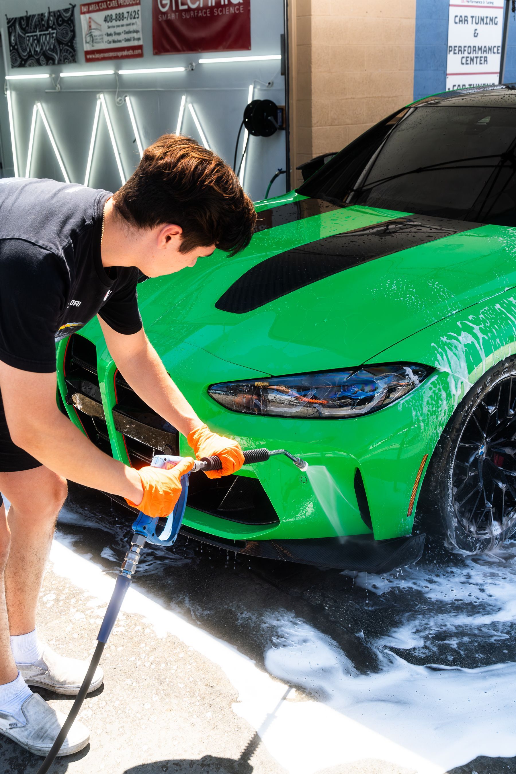 A person in orange gloves uses a pressure washer to clean a bright green sports car at a professional car wash.