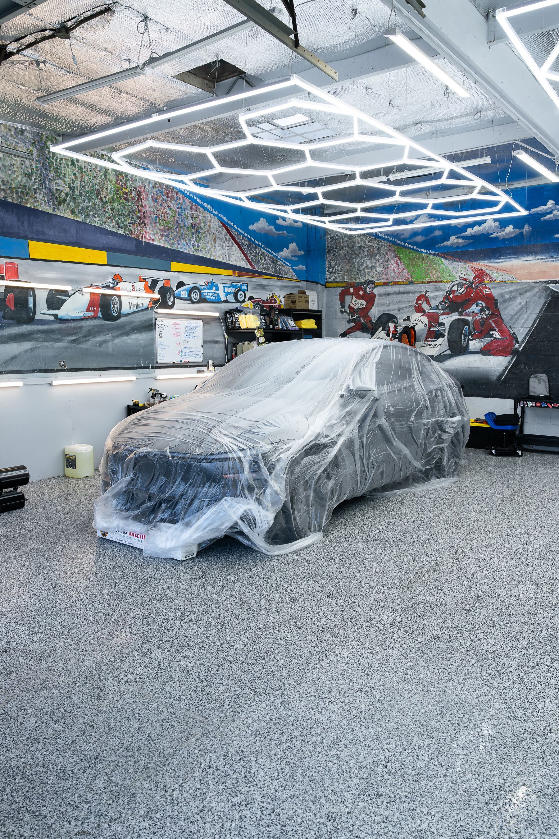 A car covered in protective plastic film inside a brightly lit garage with mural-covered walls and a speckled floor.