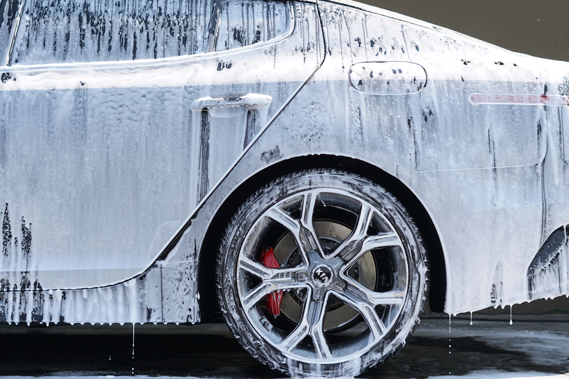 A white car covered in thick cleaning foam, focusing on the rear wheel with a red brake caliper.
