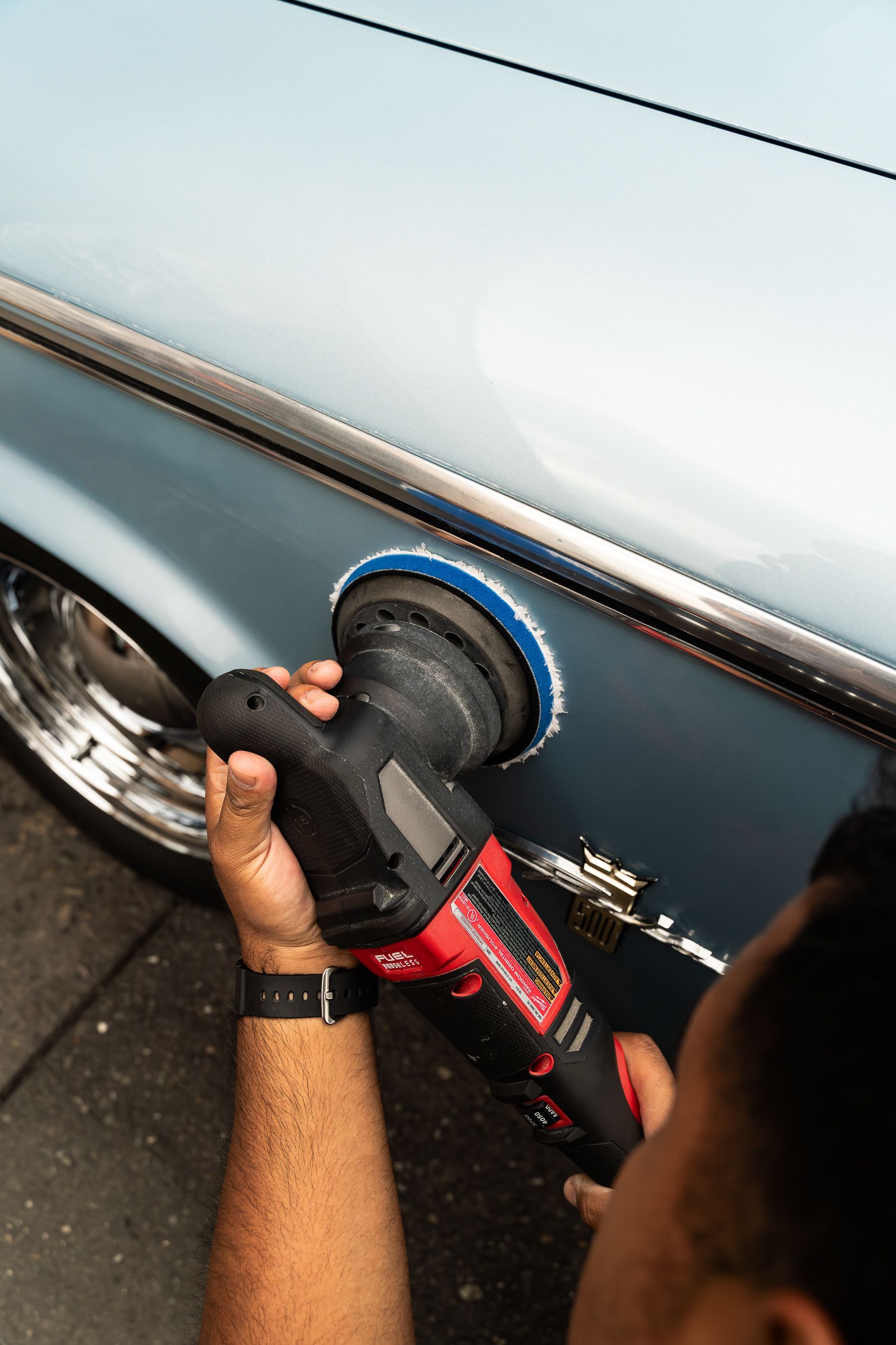 A person uses a handheld power polisher to buff the light blue side panel of a vehicle.