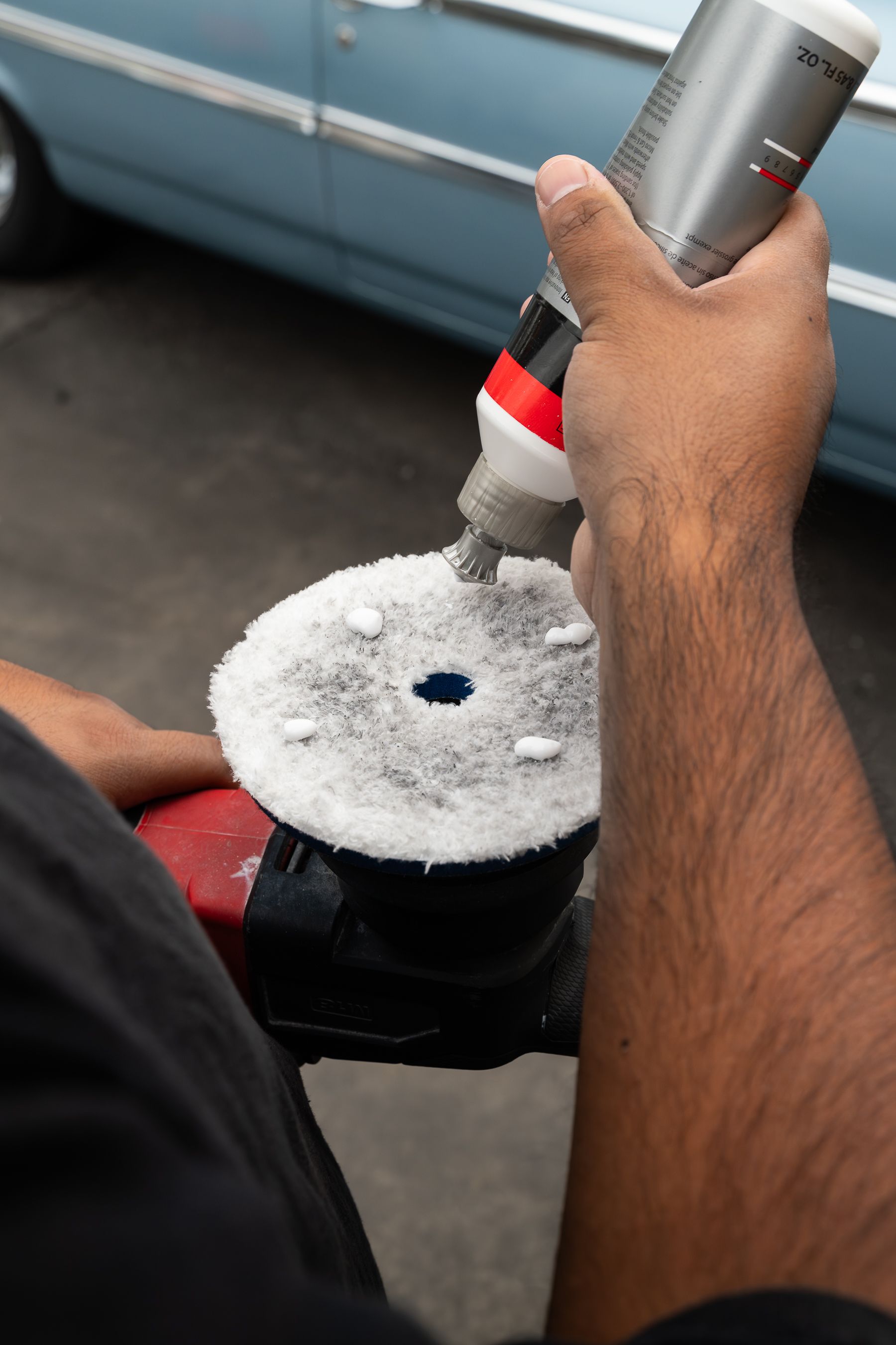 A person squeezes white polishing compound from a bottle onto the circular pad of an electric car polisher.