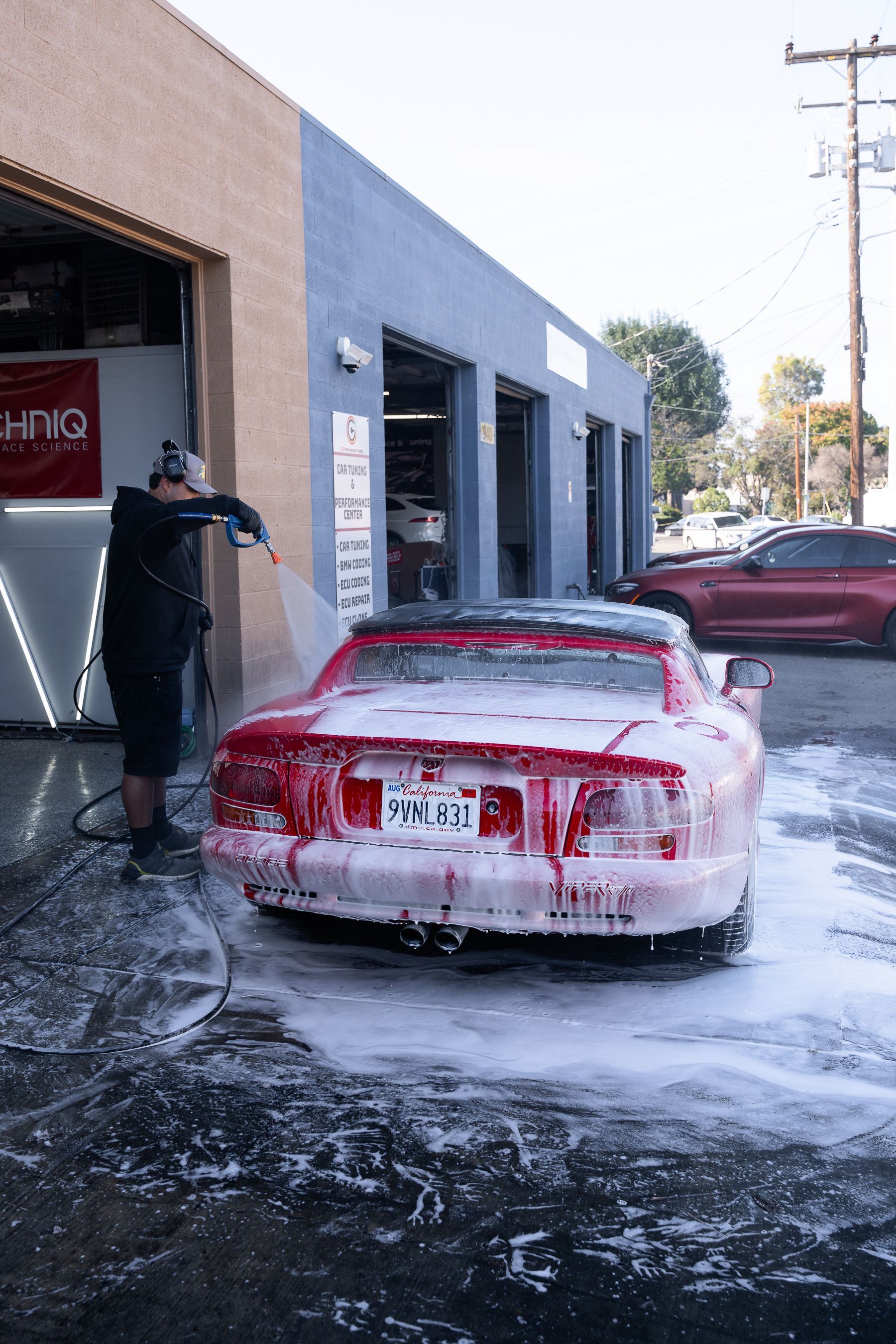 A person uses a pressure washer to rinse white foam off a red sports car parked in front of a shop.