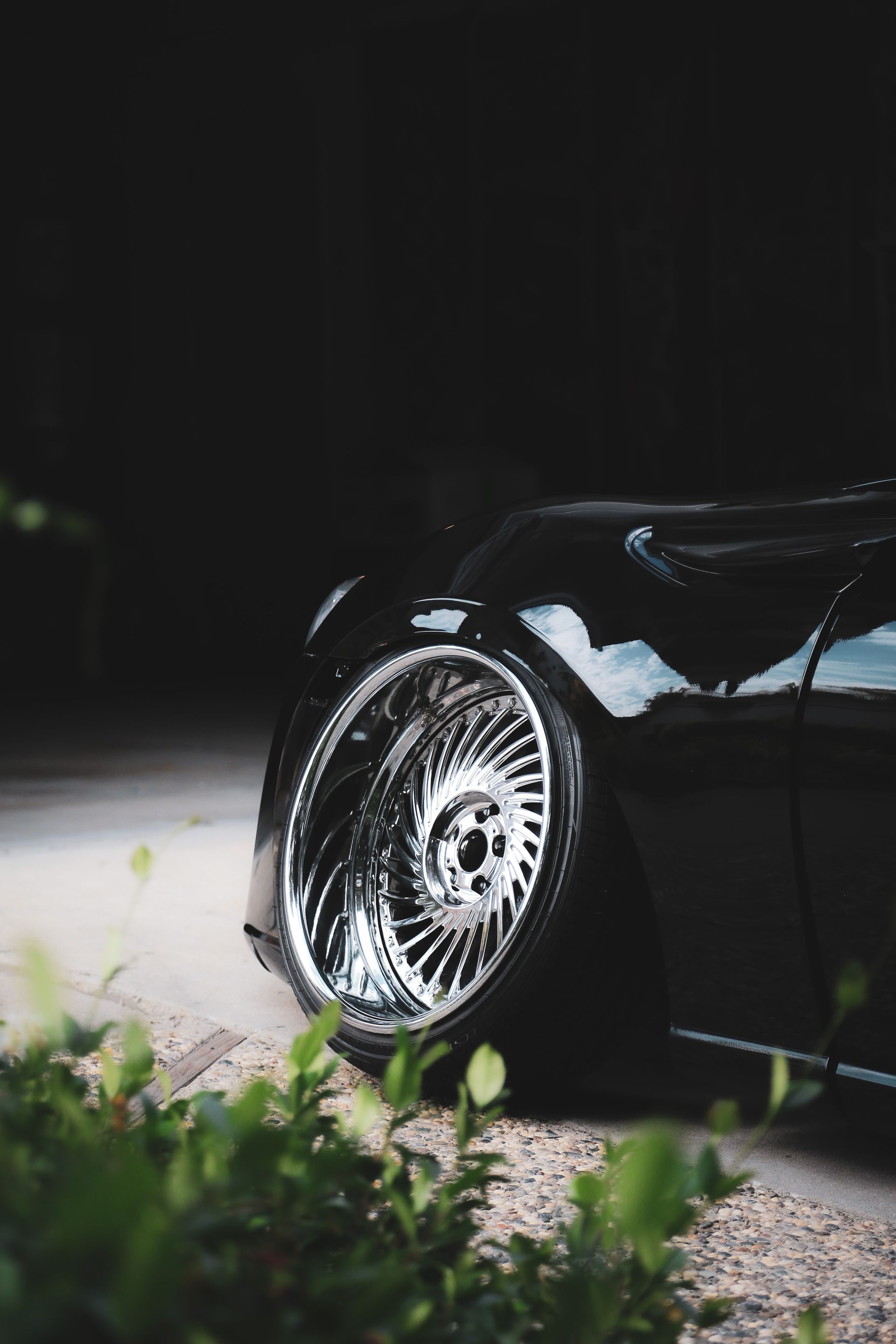 A close-up shot of a black car's shiny, deep-dish chrome custom wheel, set at a low angle near a green bush.