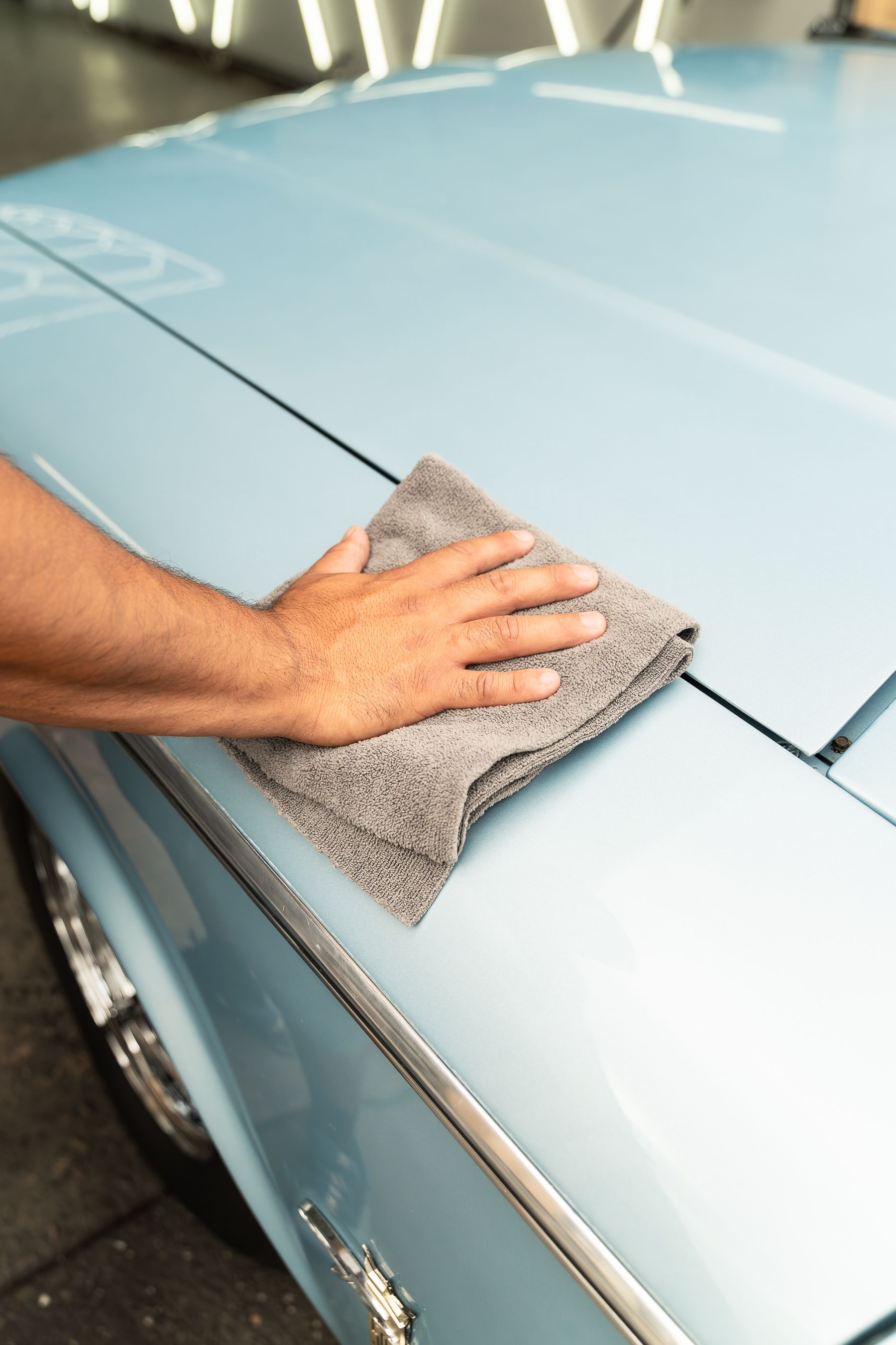 A hand uses a grey cloth to wipe the light blue hood of a car.