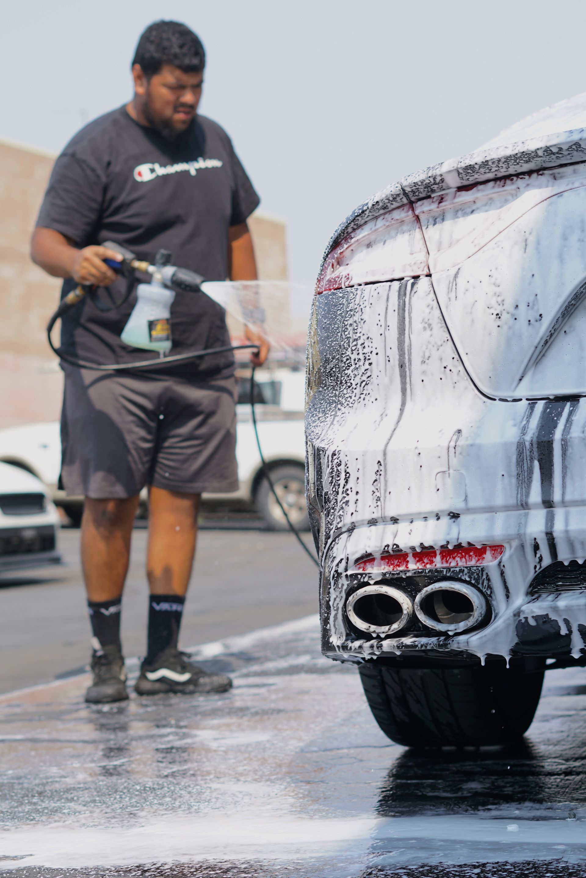 A person uses a foam cannon to apply cleaning soap to the back of a luxury car at an outdoor car wash.