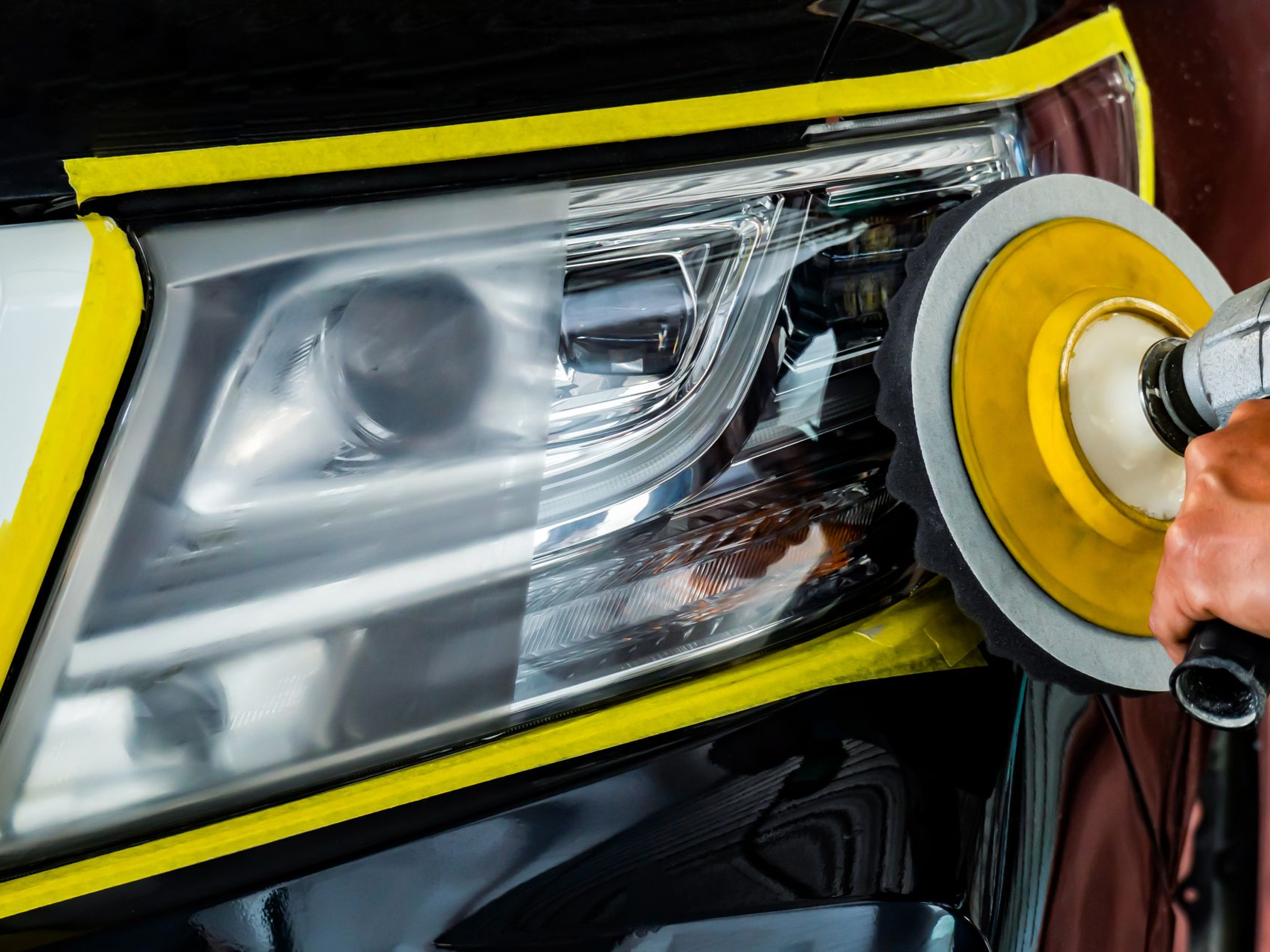 A power tool with a yellow foam pad polishes a car headlight, showing a clear contrast between clean and cloudy sections.
