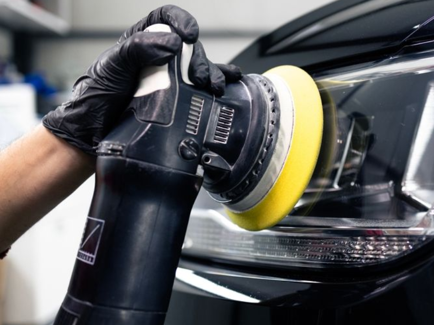 A person wearing black gloves uses a power buffer with a yellow foam pad to polish a vehicle's headlight.