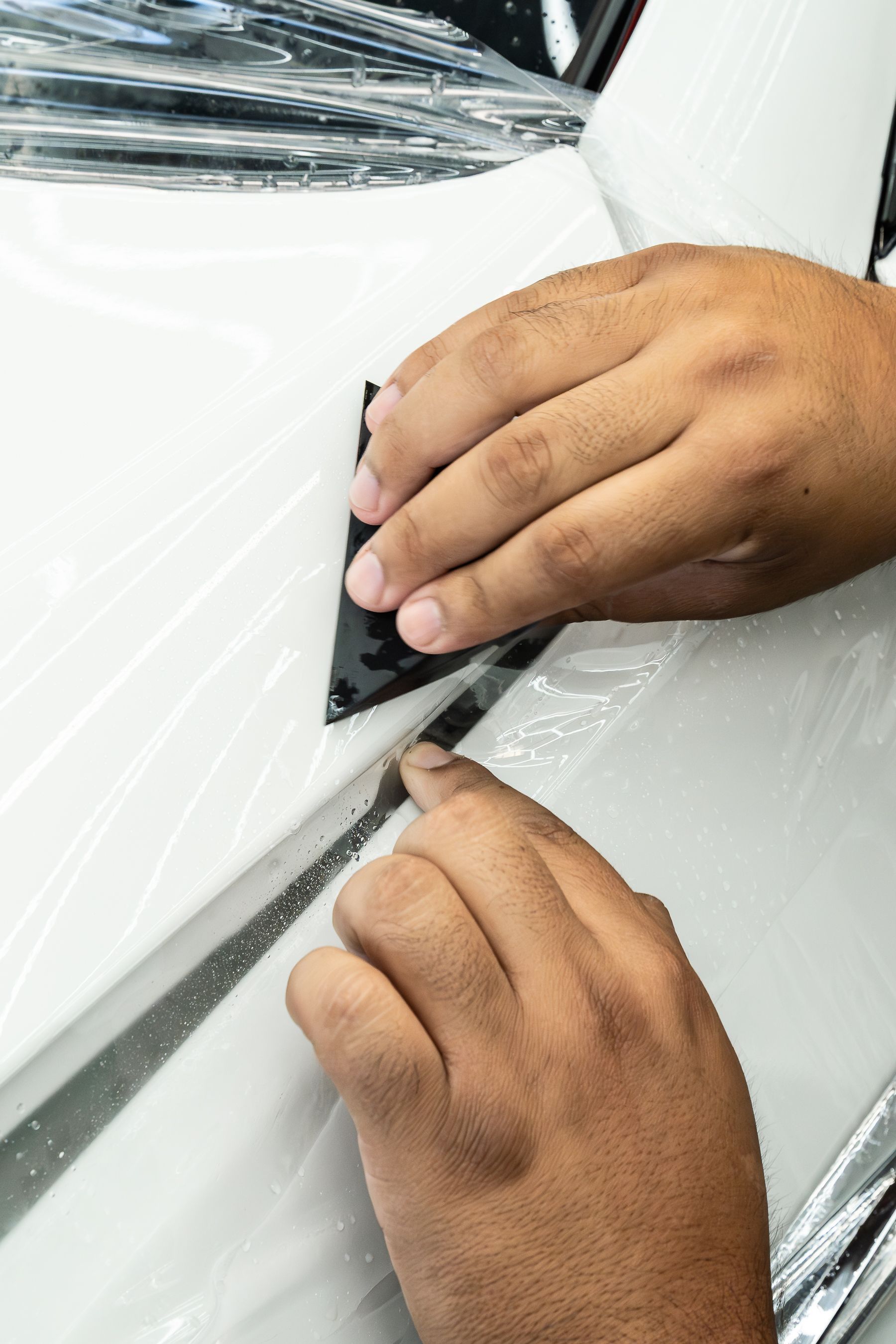 A person uses a small black squeegee tool to apply protective transparent film to a white car surface.