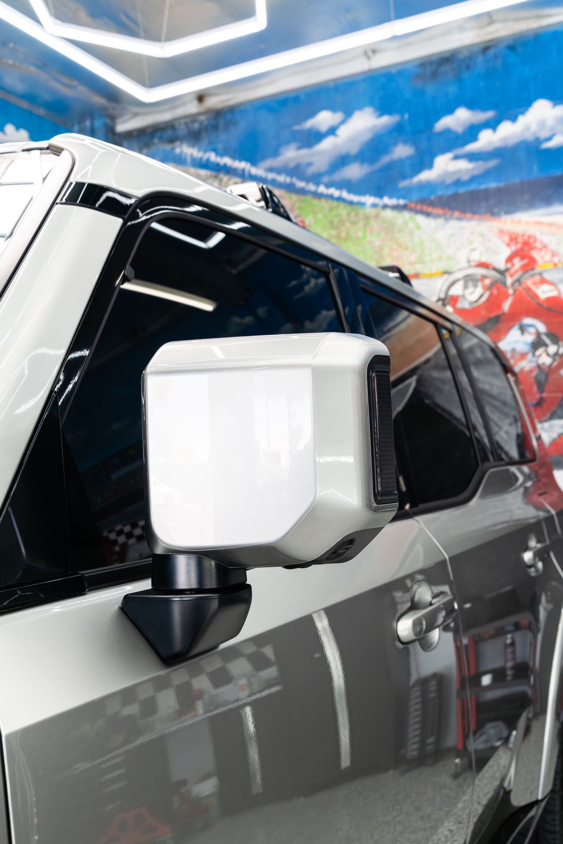 A close-up of a white car's side mirror and door, with a blurred mural featuring a red vehicle in the background.