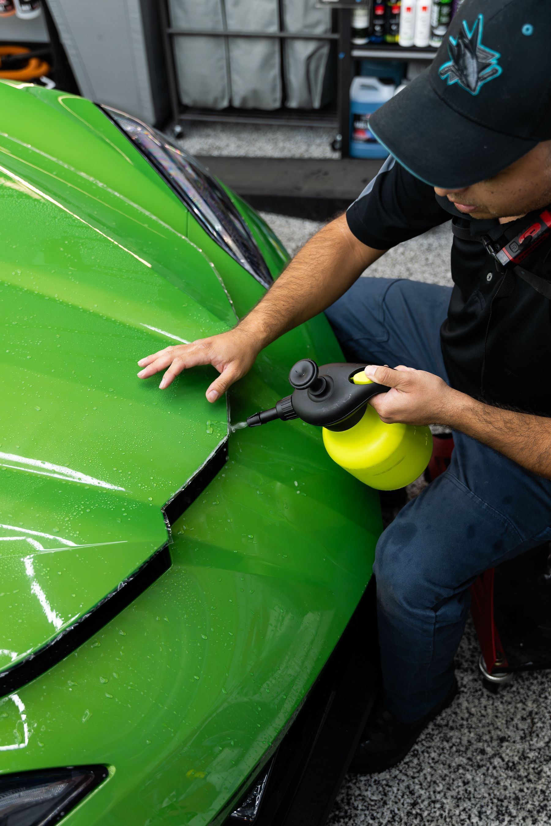 A technician in a black cap sprays a solution onto the bright green hood of a car inside a professional detailing shop.