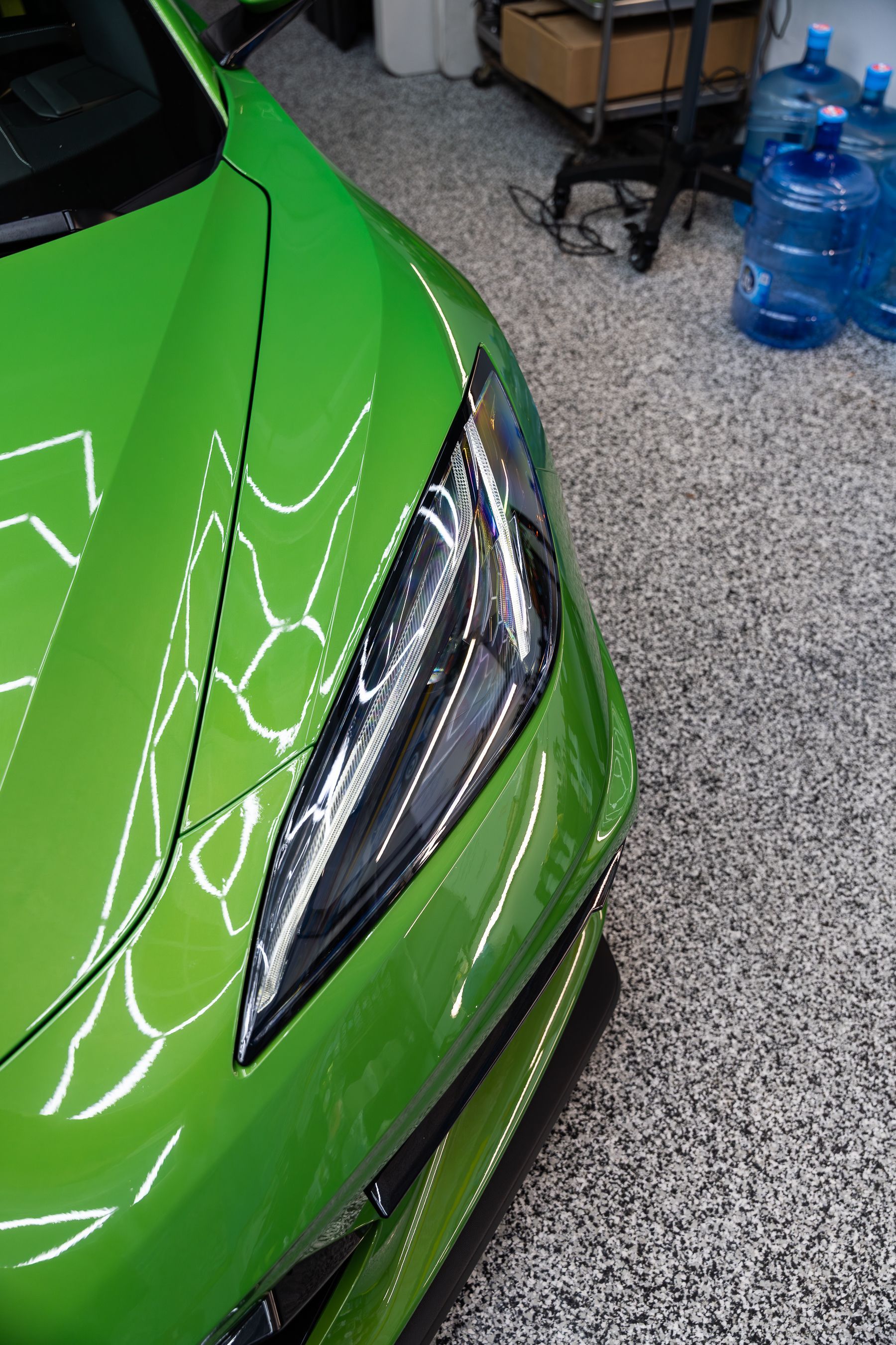 A close-up view of the front headlight and hood of a vibrant lime green sports car parked on a speckled floor.