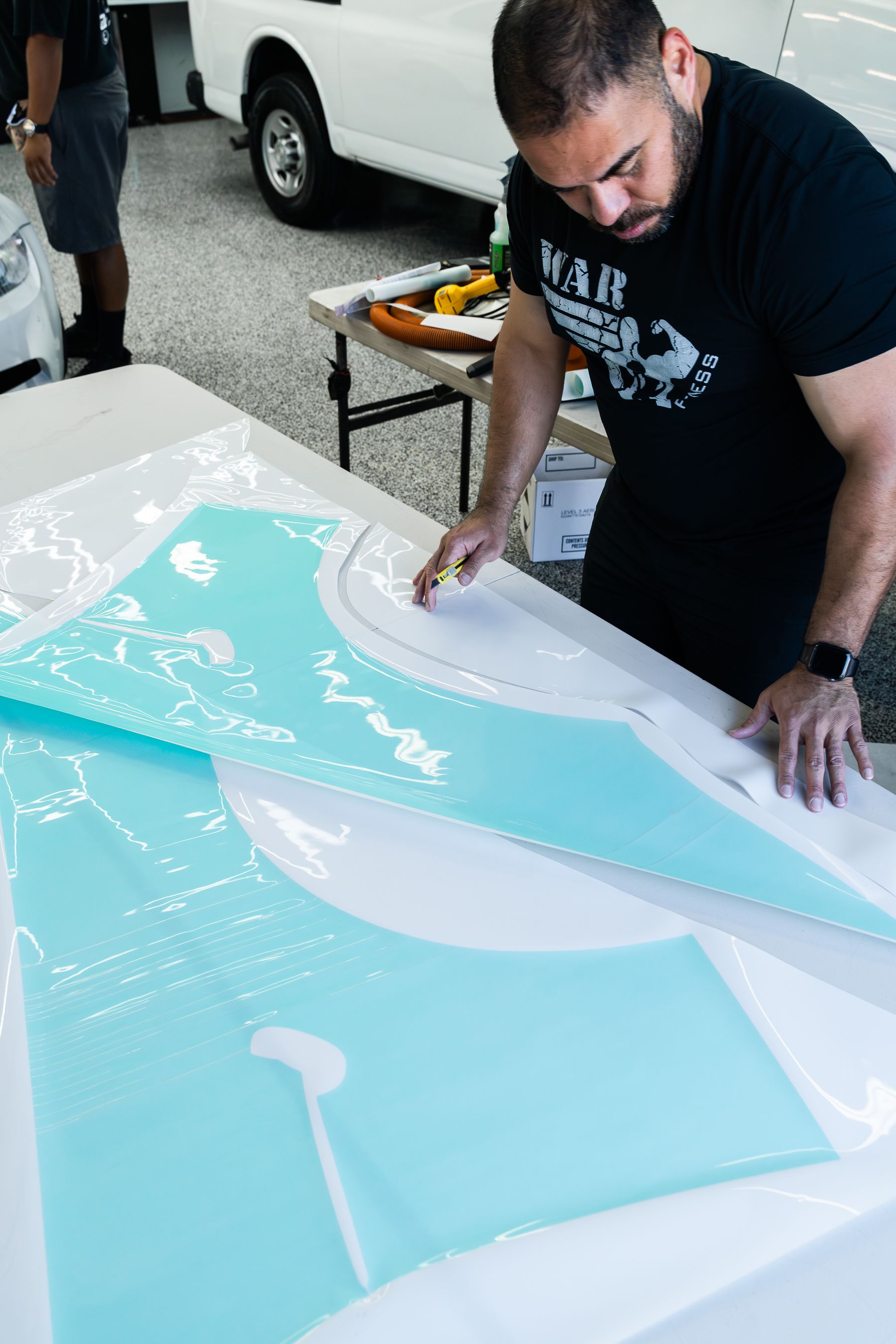 A person carefully peeling a light blue vinyl adhesive sheet from a white backing on a table in a workshop.