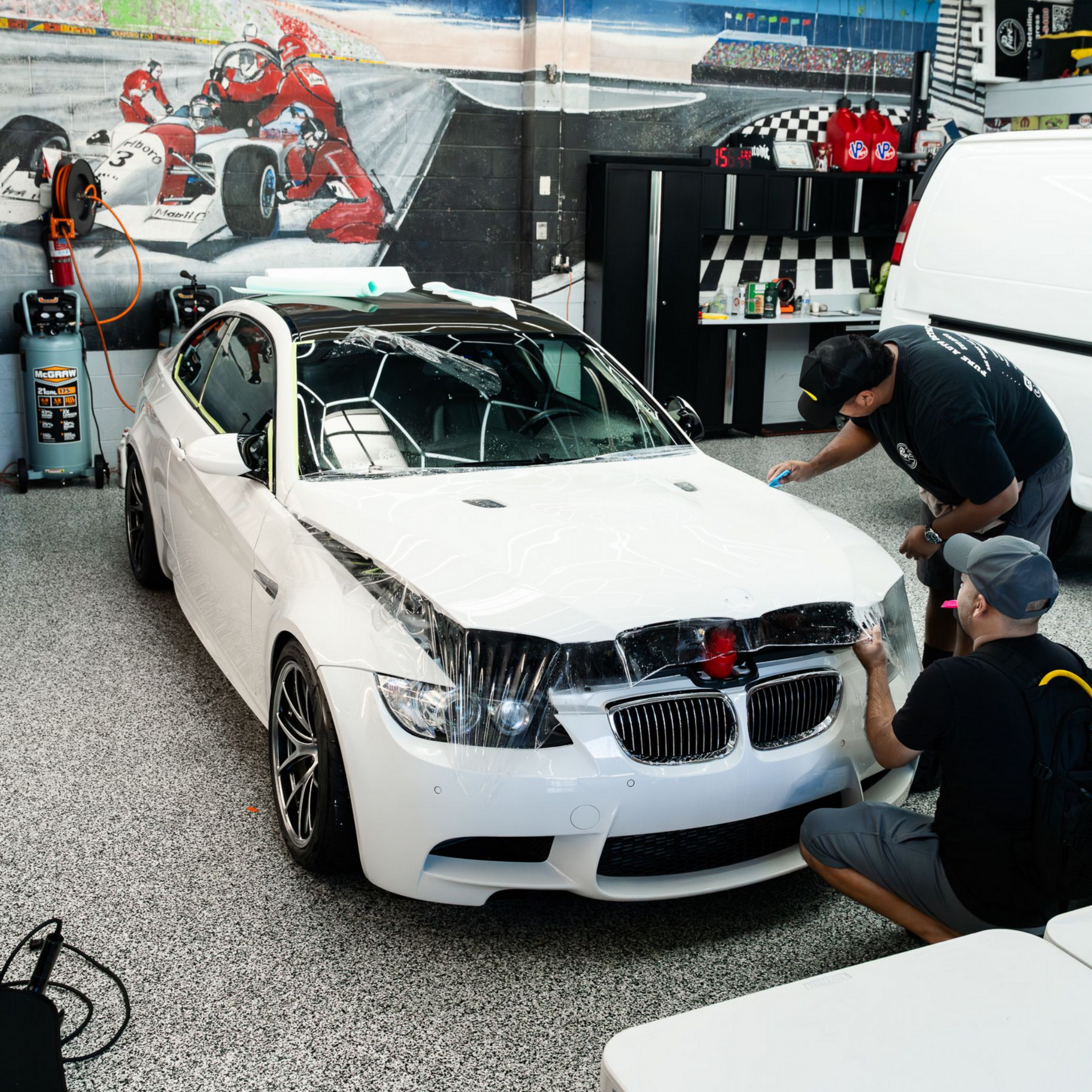 Two technicians apply clear paint protection film to the hood of a white BMW in a professional garage.