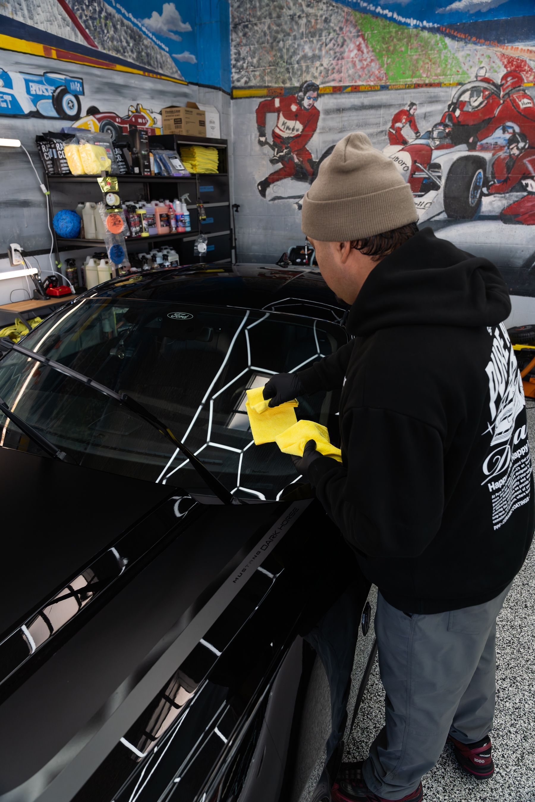 A person in a black hoodie and beanie wipes a car windshield covered in white, hexagonal vinyl patterns in a garage.