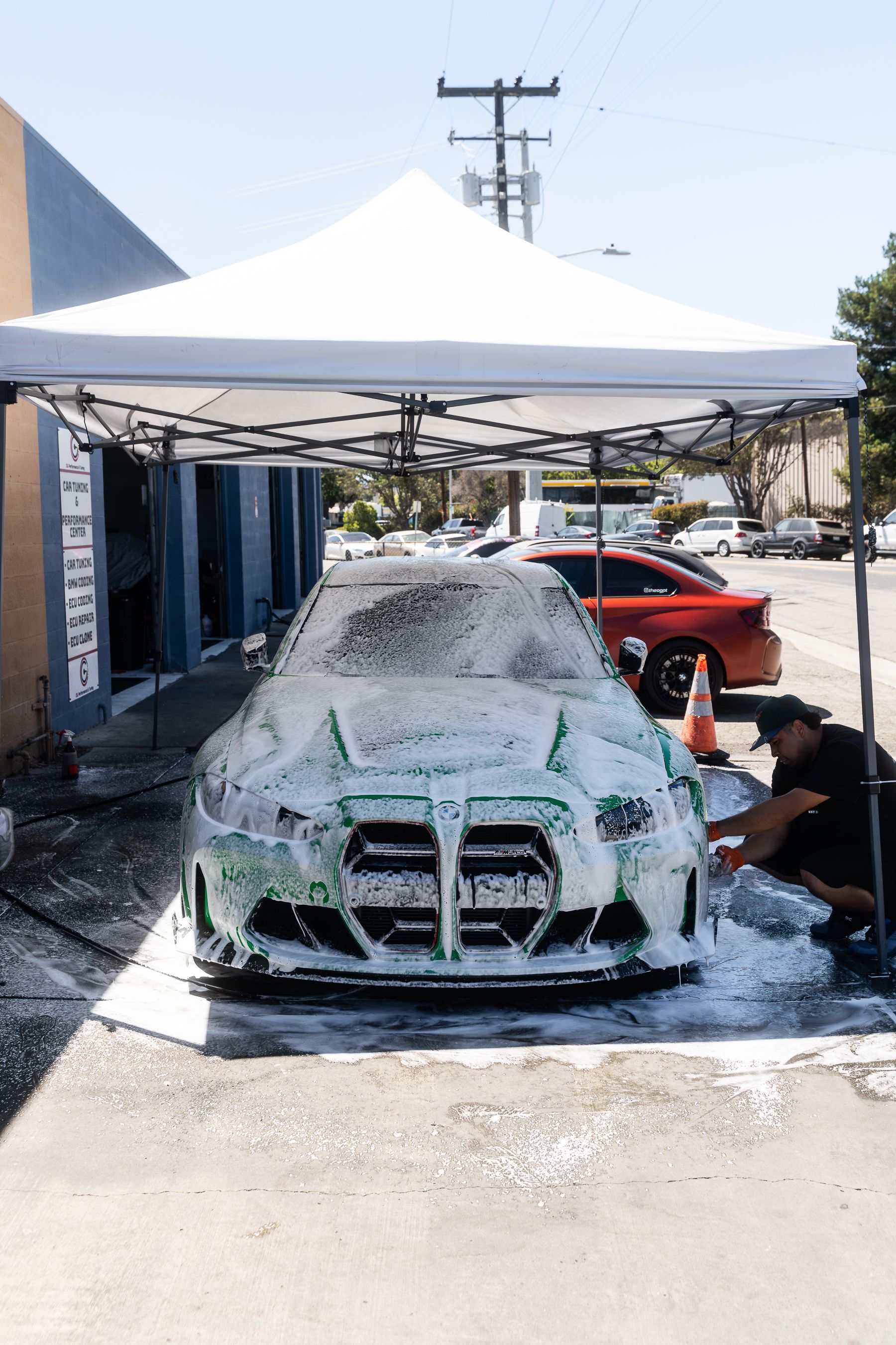 A person under a white canopy washes a car covered in thick white foam at an auto detailing shop.