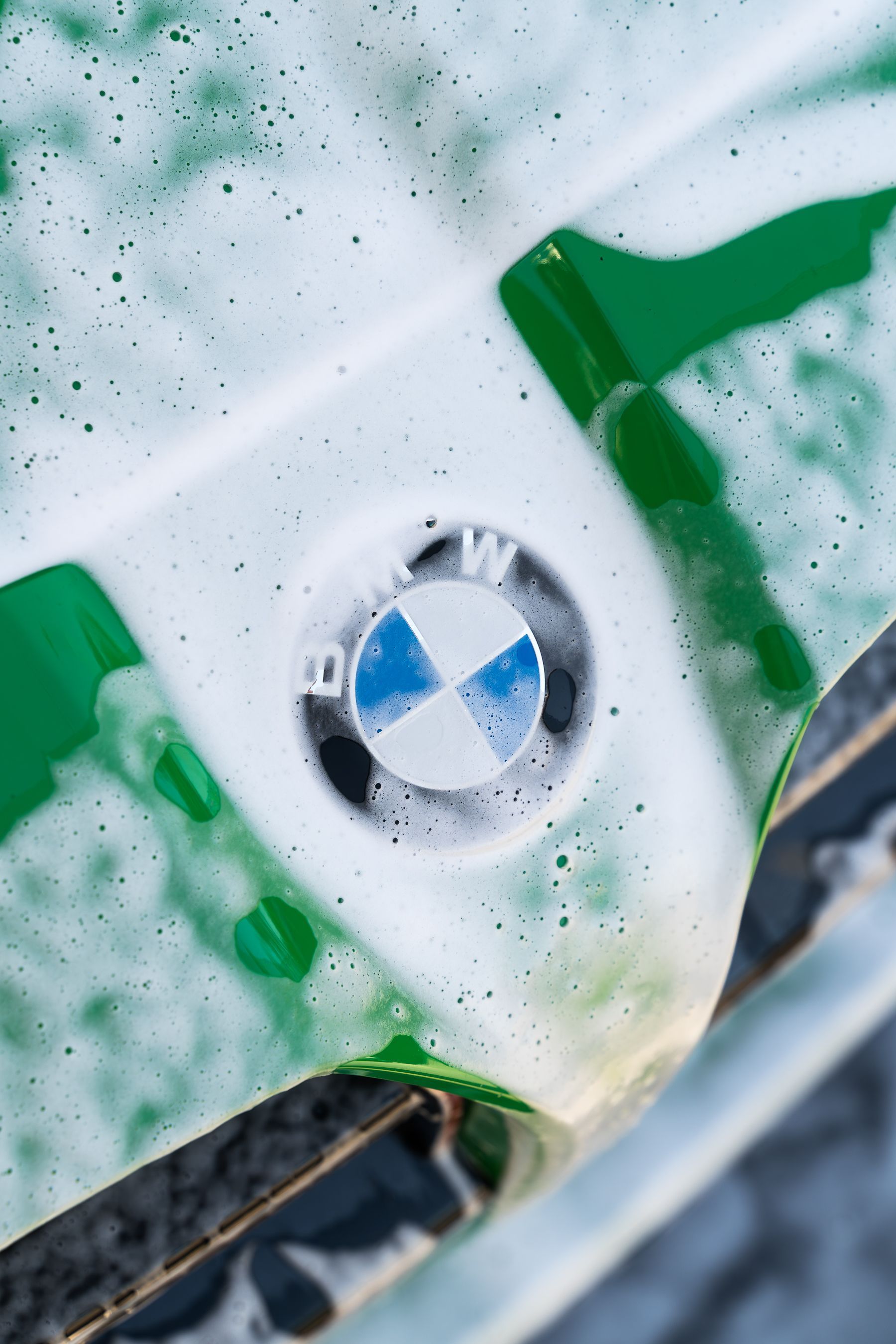 A BMW logo on a green car hood, covered in thick white cleaning foam during a car wash.