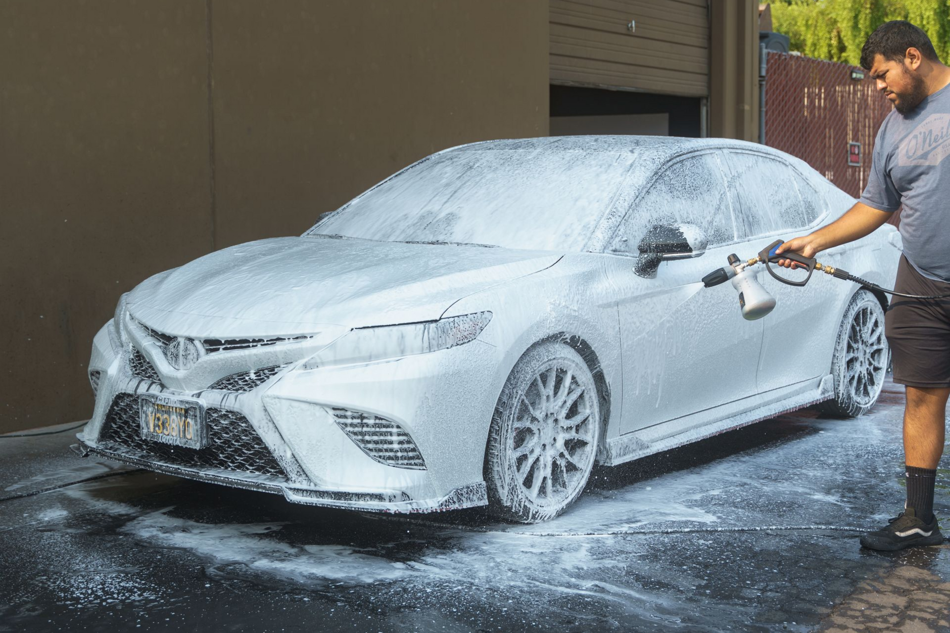 A person in a gray shirt uses a foam cannon to apply thick white soap to a white sedan parked on a driveway.