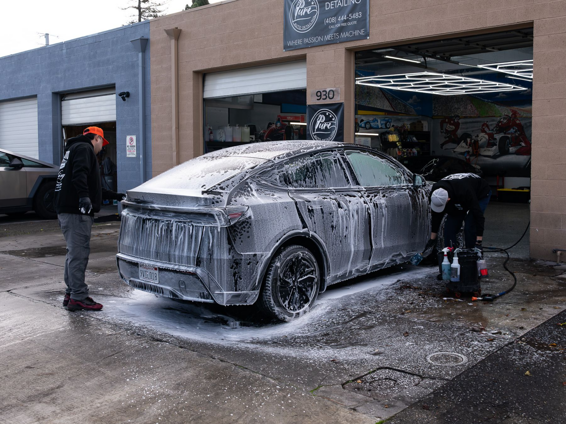 Two technicians wash a dark SUV covered in thick white soap suds outside a garage bay.