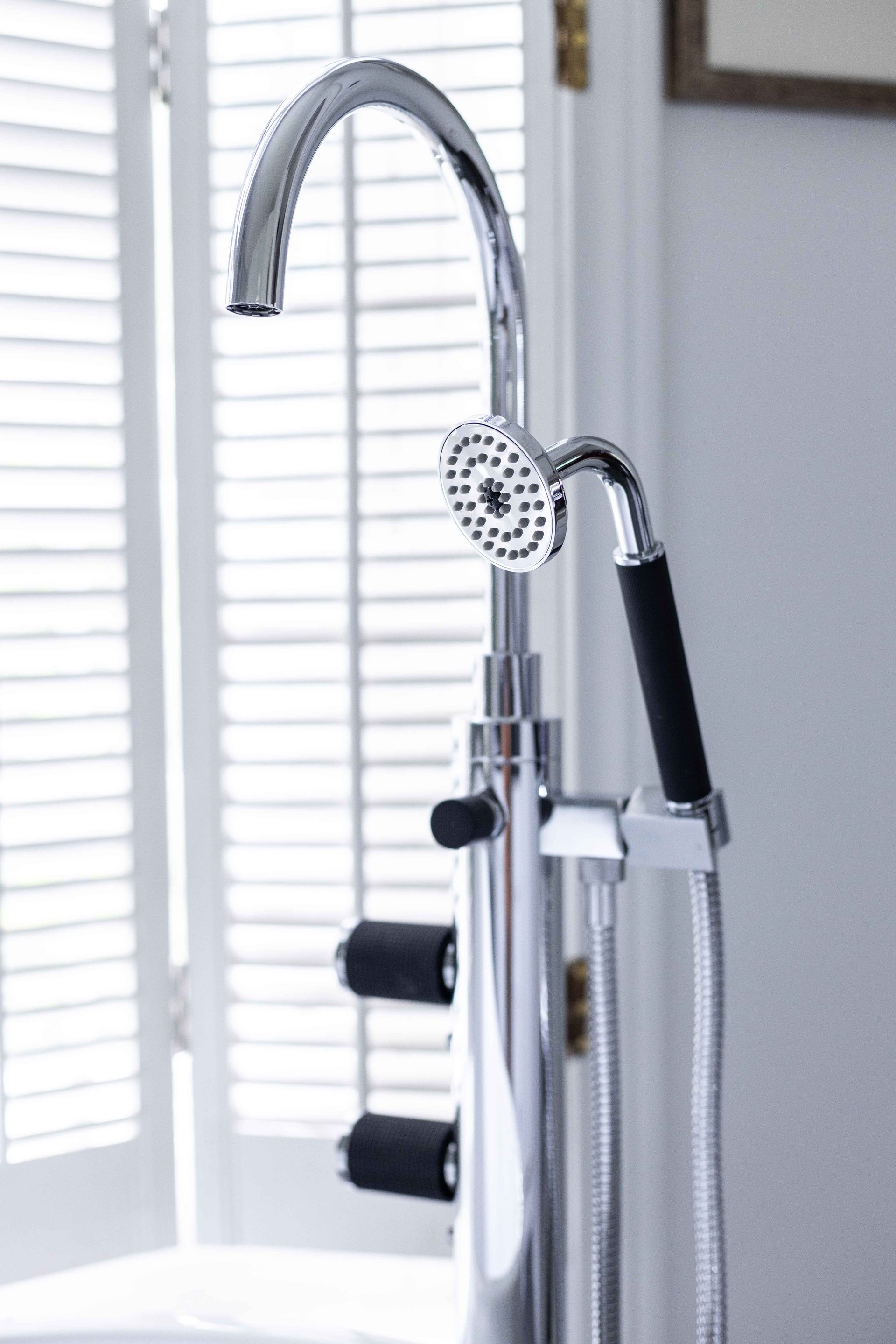 Robinet de baignoire sur pied chromé avec douchette, près d'une fenêtre à stores.